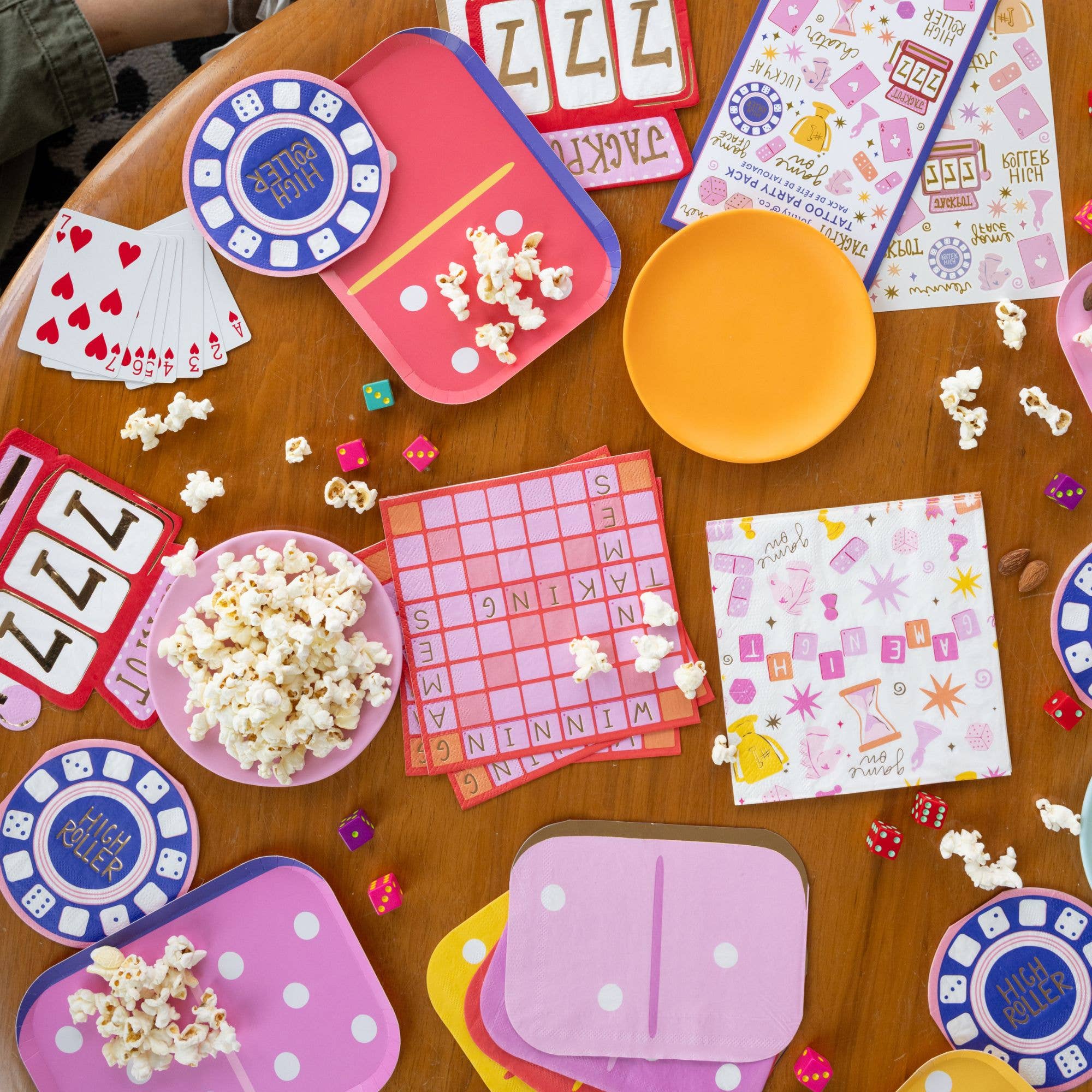 Overhead game night table scene with game night dinner plate, popcorn, playing cards, scrabble board napkins, High Roller poker chip napkins, dice, and jackpot slot machine plates on a wood table
