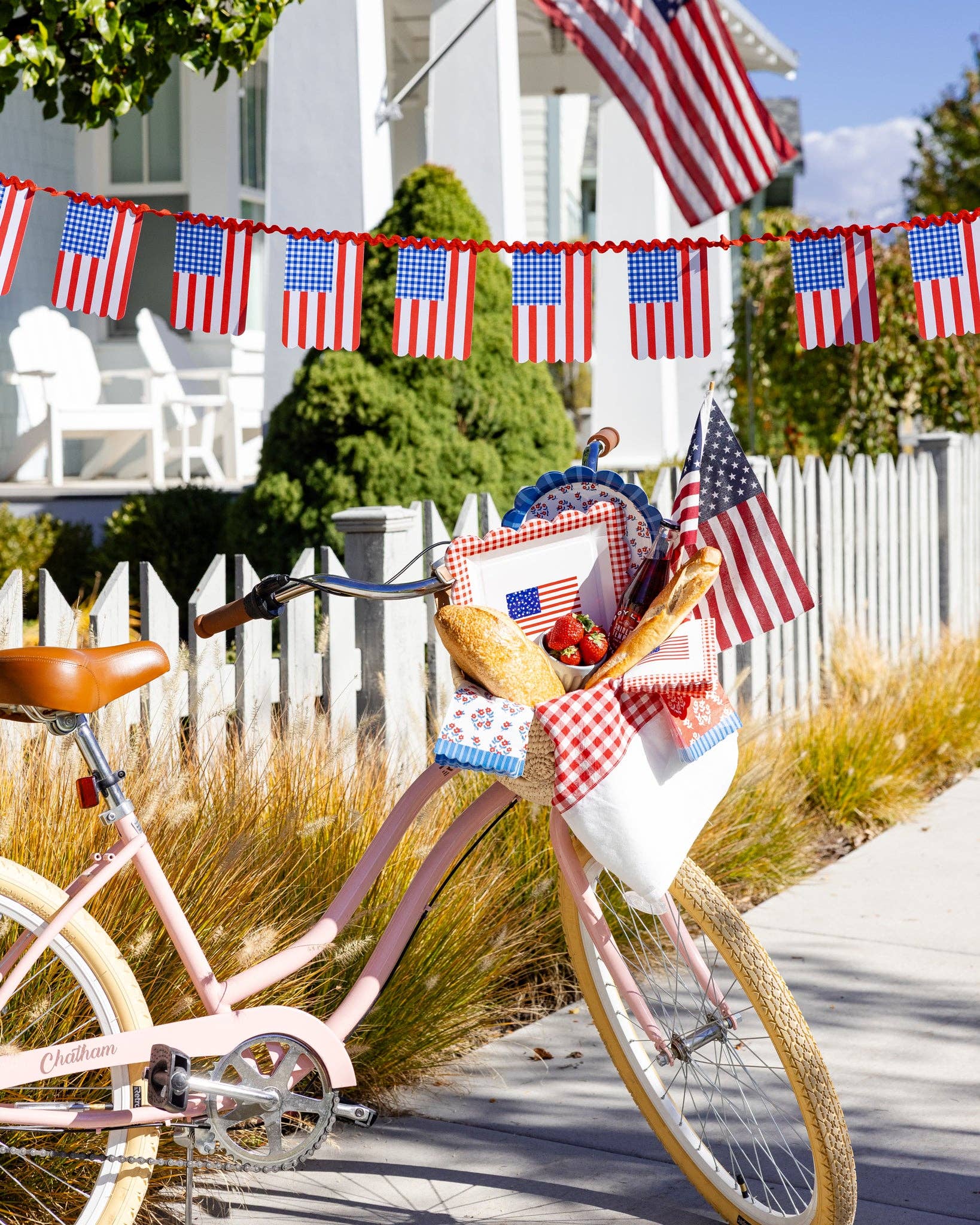 Pink bicycle with basket filled with gingham flag paper plates, floral napkins, baguettes, and American flags in front of white picket fence with patriotic bunting