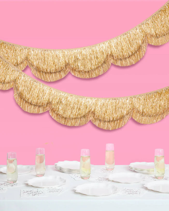 Double-layered gold scalloped fringe banner hanging in two tiers on pink background above styled bridal table with champagne flutes and white plates