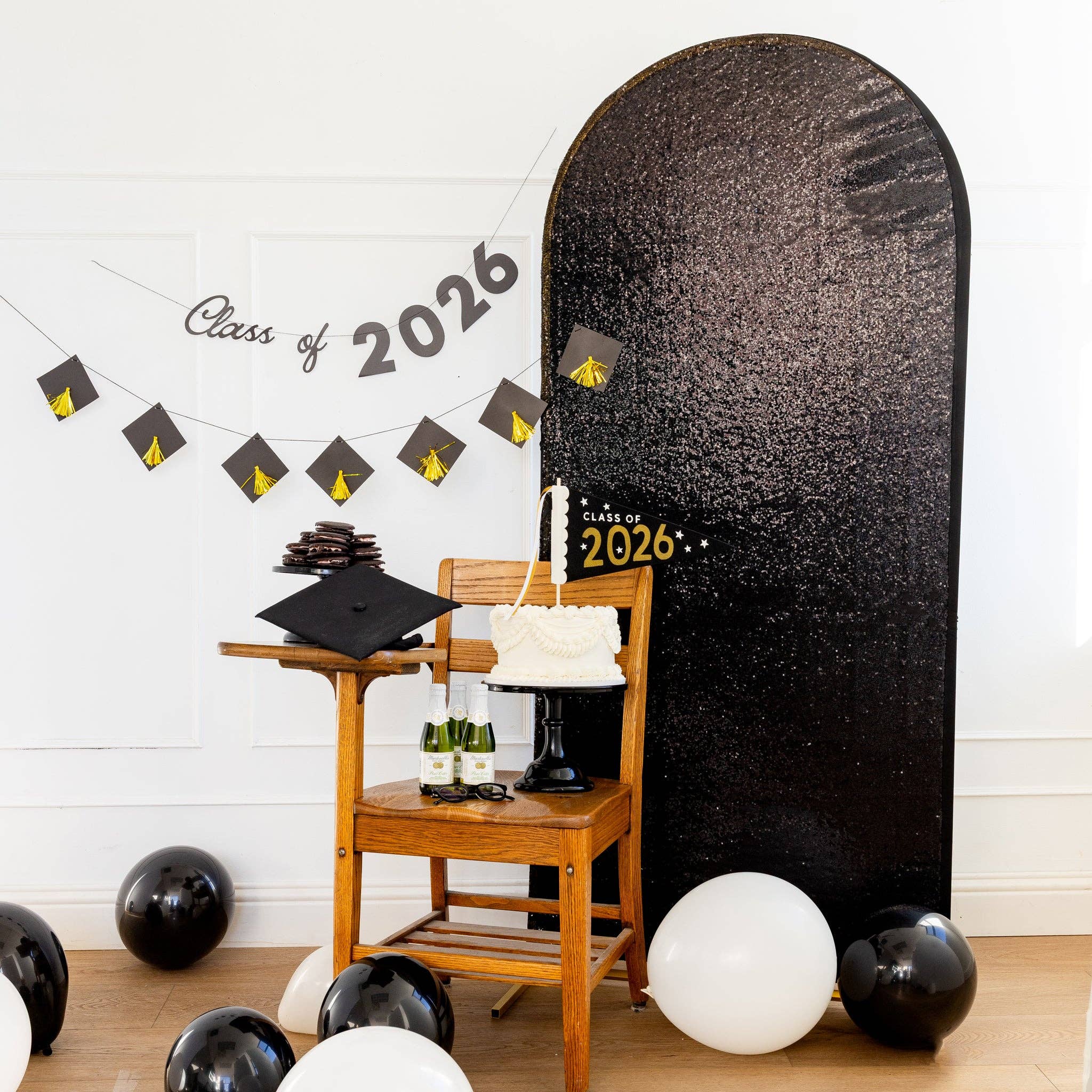 A Class of 2026 script banner and graduation cap banner hung as a photo wall backdrop next to a black sequin arch.