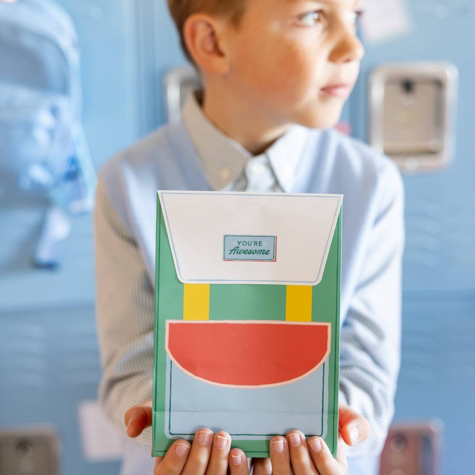 Child holding green version of backpack-shaped treat bag in classroom setting.