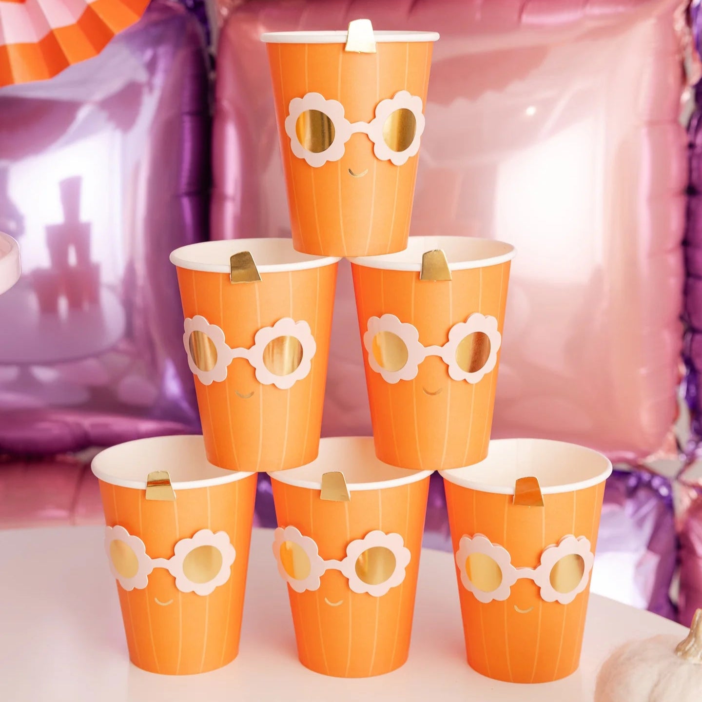 Pyramid of Groovy Pumpkin cups on party table; metallic sunglasses design and gold stems pop against pink-purple balloon wall and striped fans.