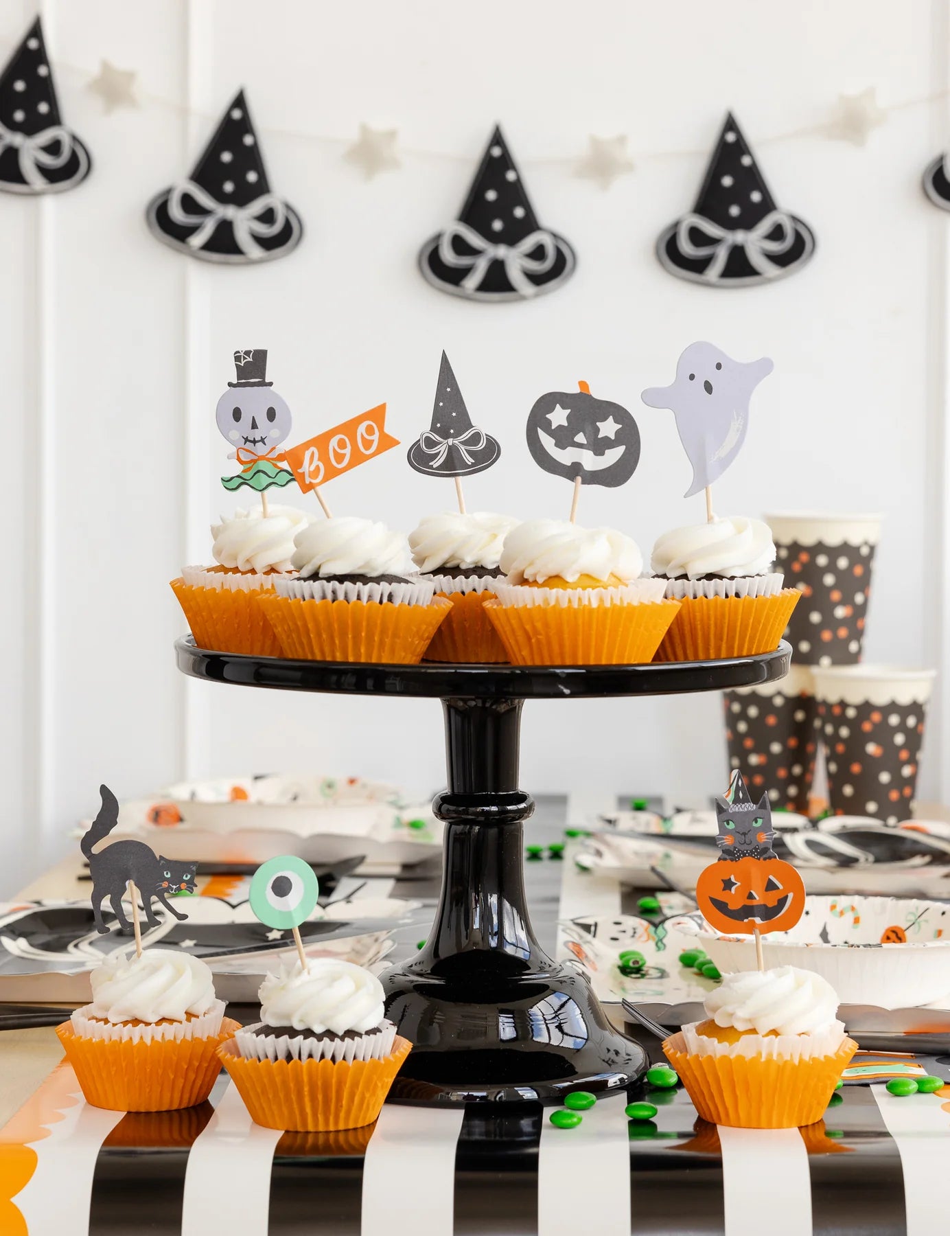 Close-up of cupcakes on black stand topped with Halloween picks; witch-hat banner blurred in background.