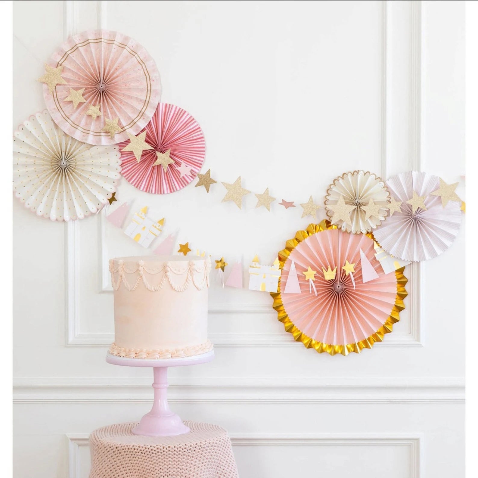 White paneled wall decorated with pastel paper fan rosettes, glitter-gold star garland, and a pink-and-gold princess icon banner above a blush layer cake on a pale lavender pedestal stand.