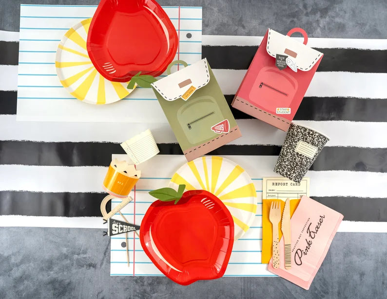 Overhead view of two red apple plates, yellow sunburst plates, and mini backpack favor boxes displayed on notebook‑style placemats atop a bold black‑and‑white striped runner.