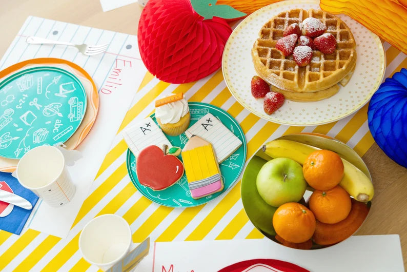 Breakfast spread on a table runner: teal doodle plates, pencil‑shaped cookies, honeycomb apple decorations, waffles with strawberries, and loose‑leaf placemats layered below.