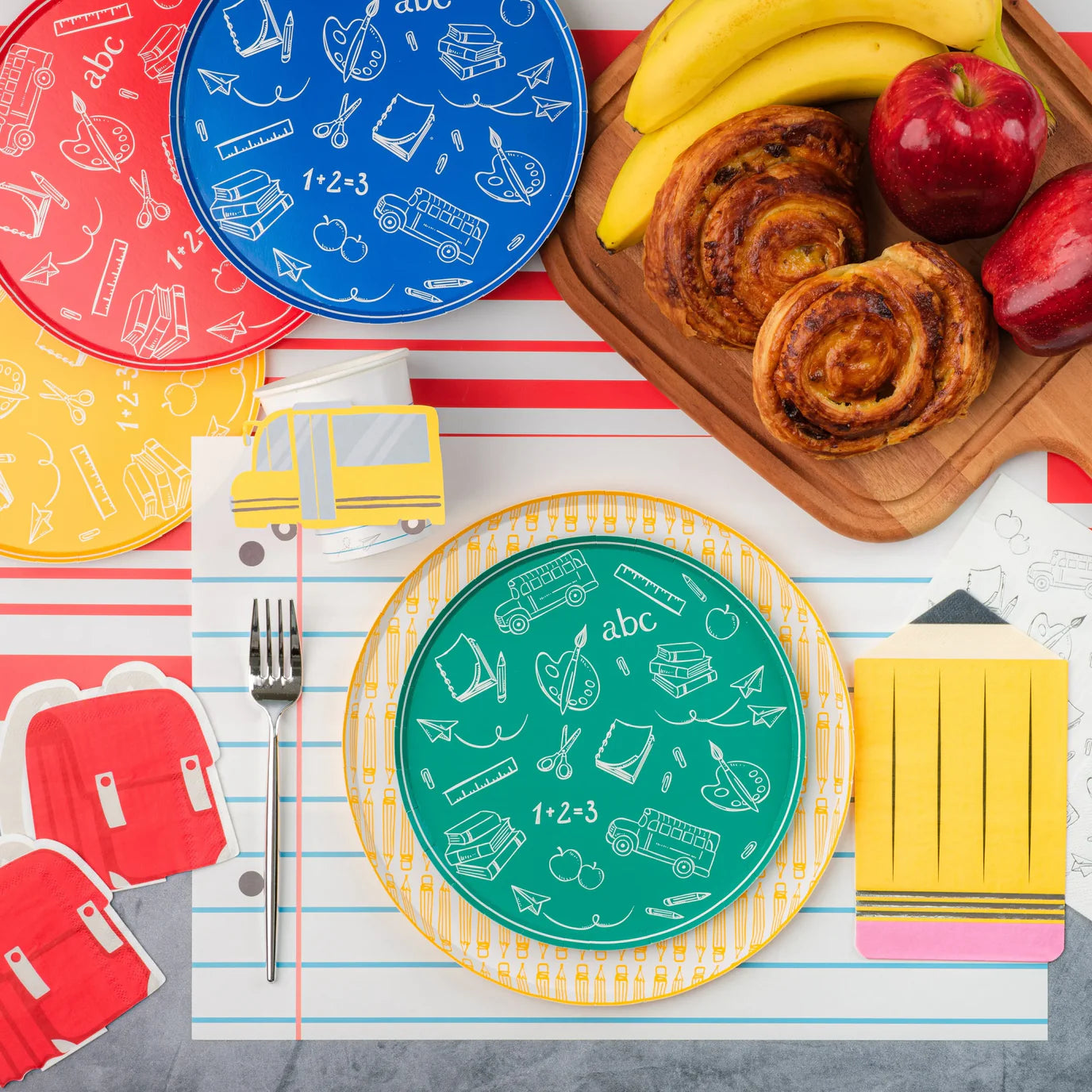 Bright breakfast spread with pastries, bananas, green school‑icon plate on pencil dinner plate, and pencil napkin