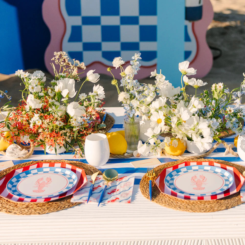 Overhead view of nautical summer party table with lobster checkered plates, lobster napkins, rattan chargers, and white floral centerpieces on blue striped runner