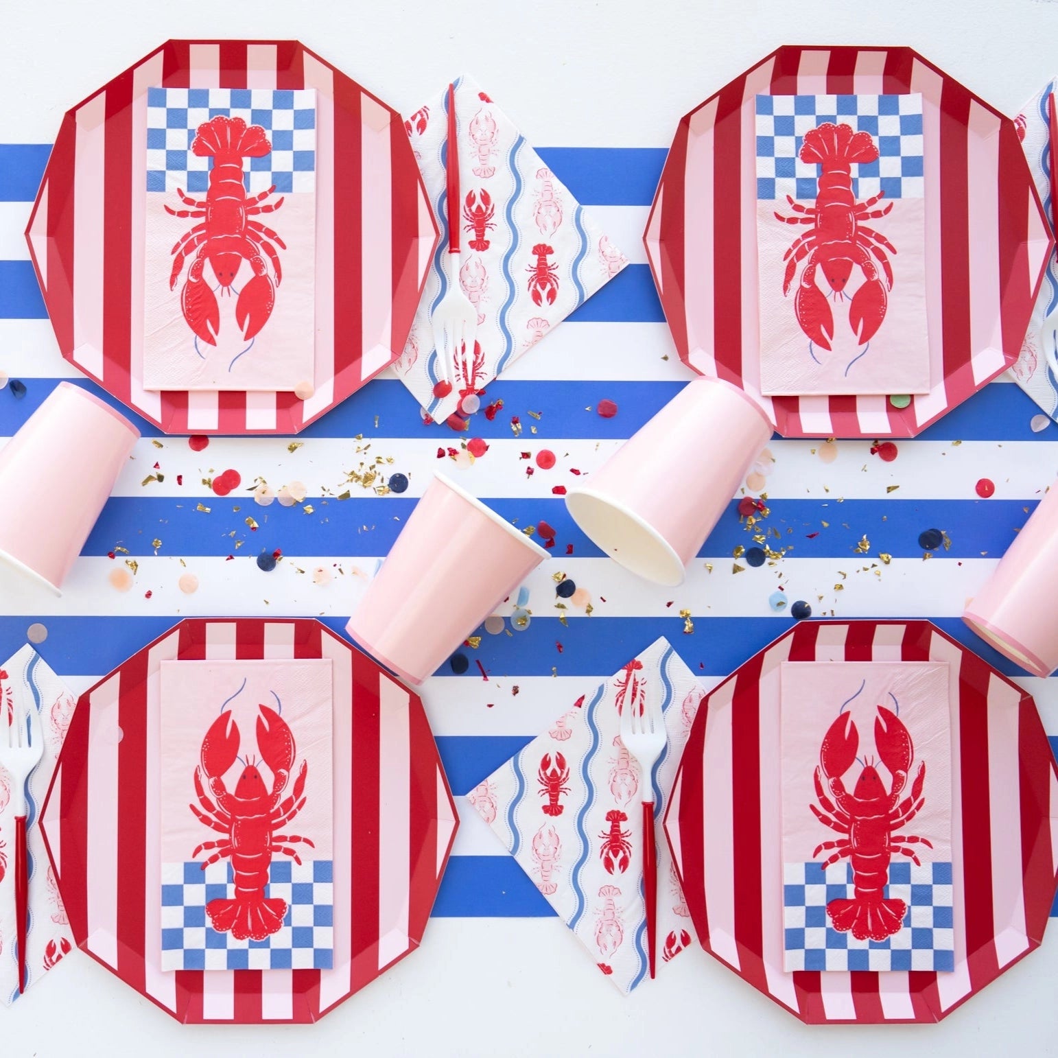 Nautical tablescape with striped red plates, lobster lunch napkins, pink cups and blue‑white runner scattered with confetti.