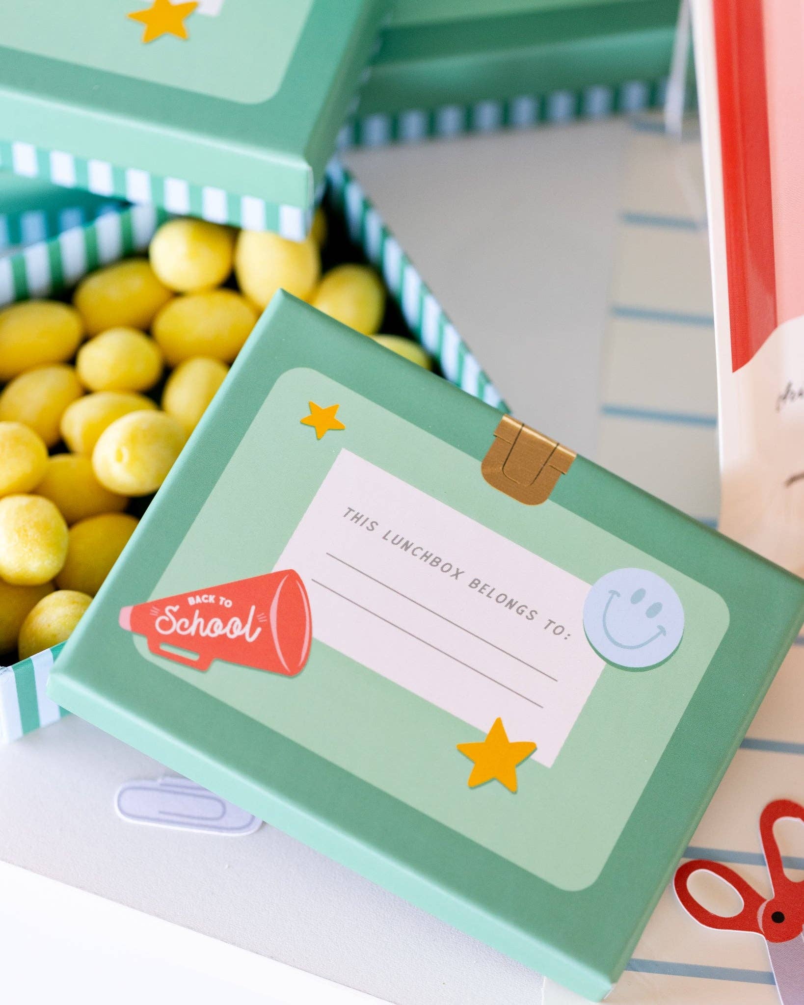 Close-up of teal lunchbox shaped treat box with gold latch, back-to-school megaphone icon, yellow stars, and smiley face sticker detail, filled with yellow candies
