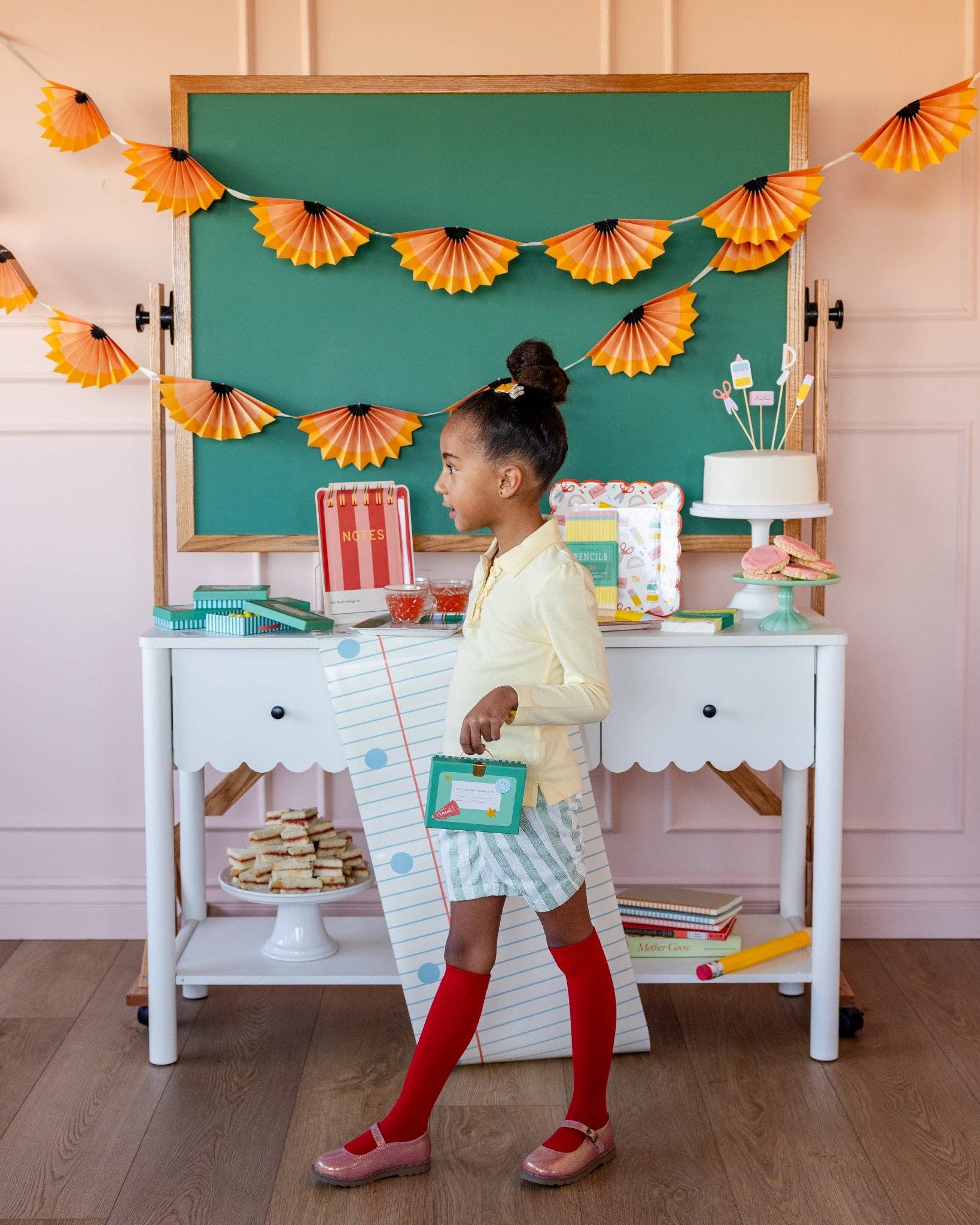 Young girl in yellow cardigan holding small teal lunchbox shaped treat box at styled back to school party with pencil shaving banner backdrop