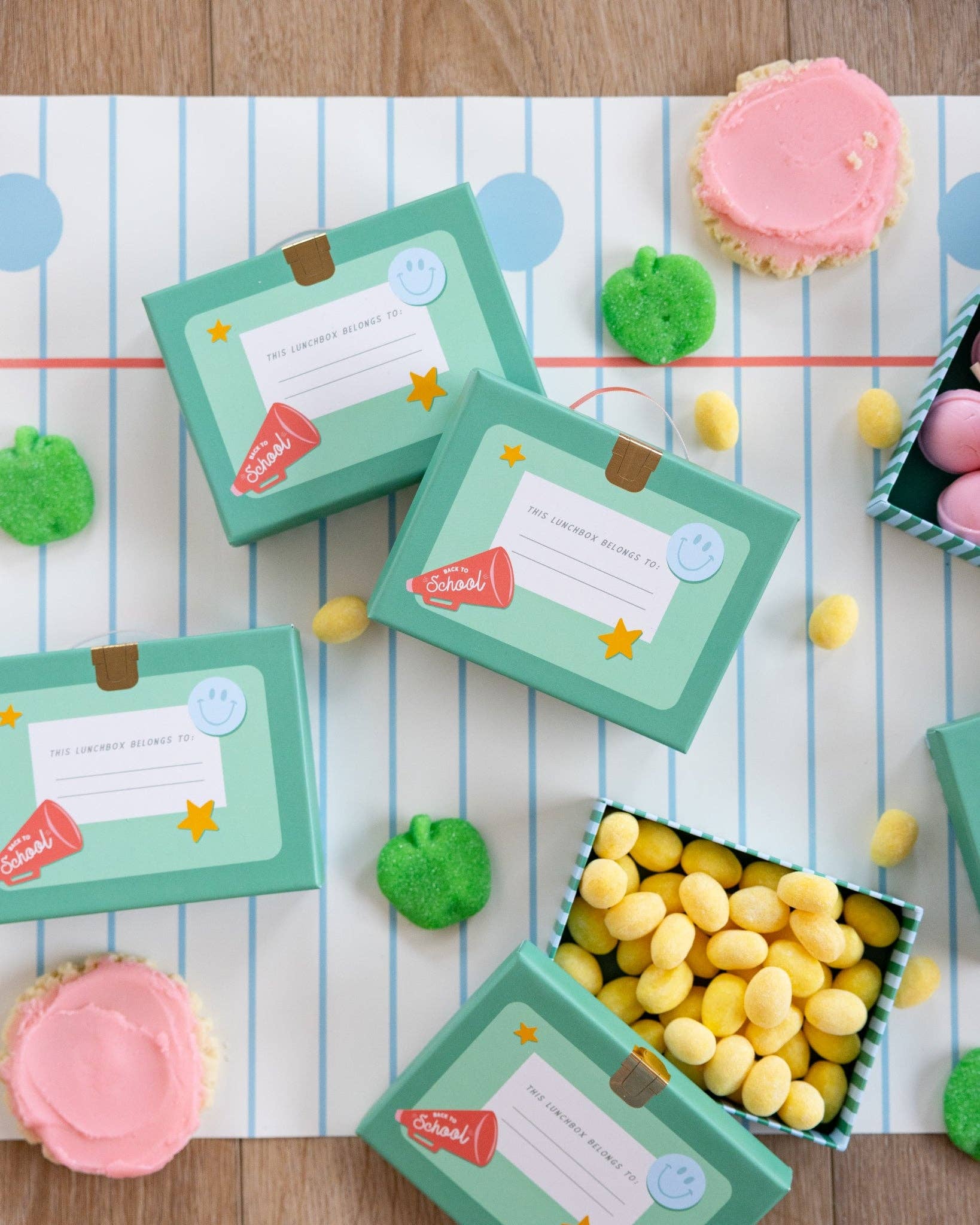 Flatlay of teal lunchbox shaped treat boxes on notebook paper background surrounded by sugar cookies, green apple candies, and yellow jelly candies