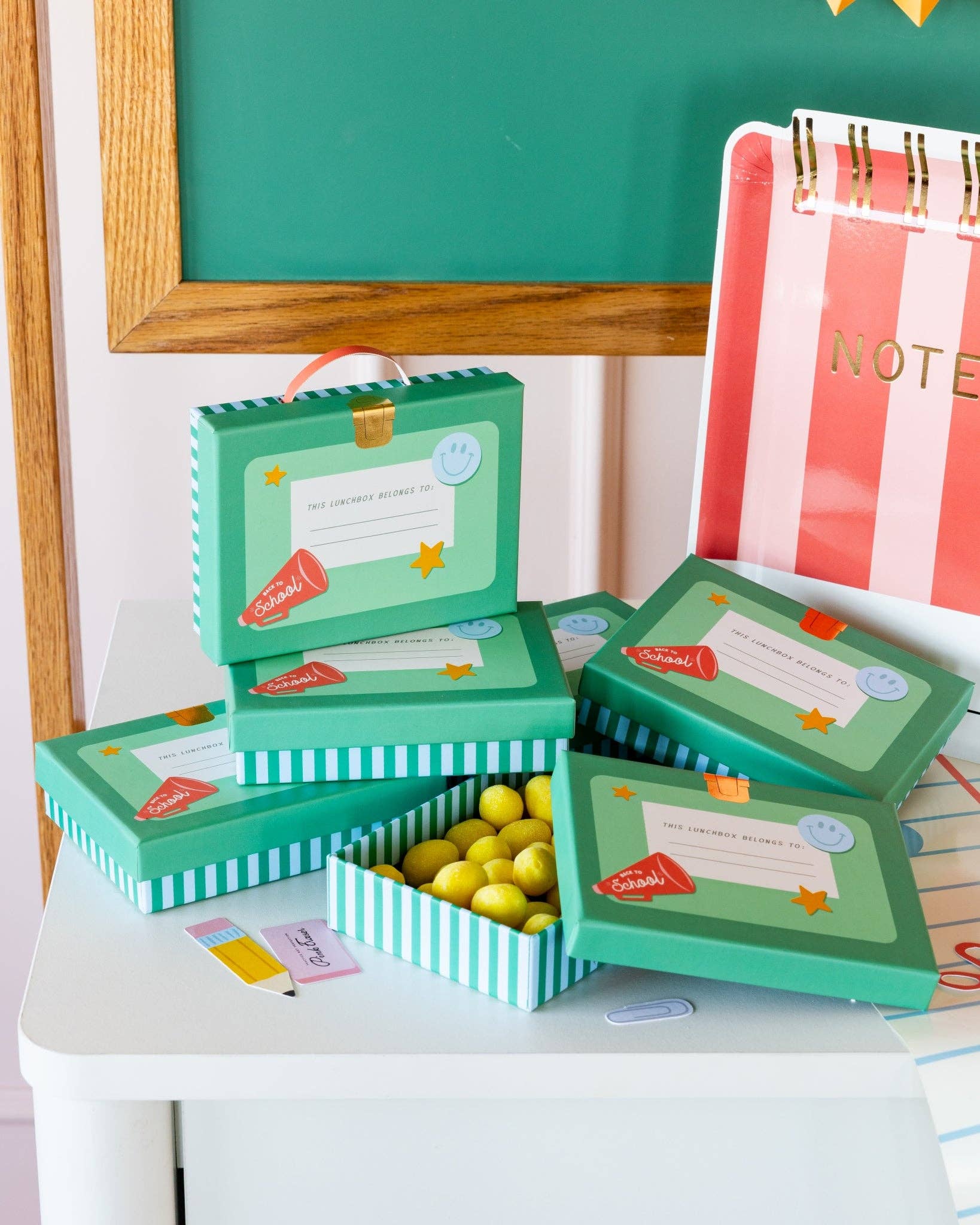 Stack of teal lunchbox shaped treat boxes on white desk with chalkboard backdrop, one open to show striped interior and yellow candies