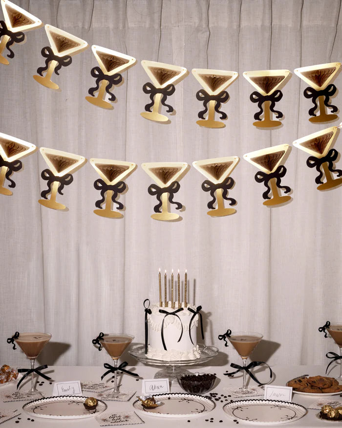 Two rows of martini glass bow banners hanging above an espresso martini birthday table with cake, chocolates, place cards, and cocktails
