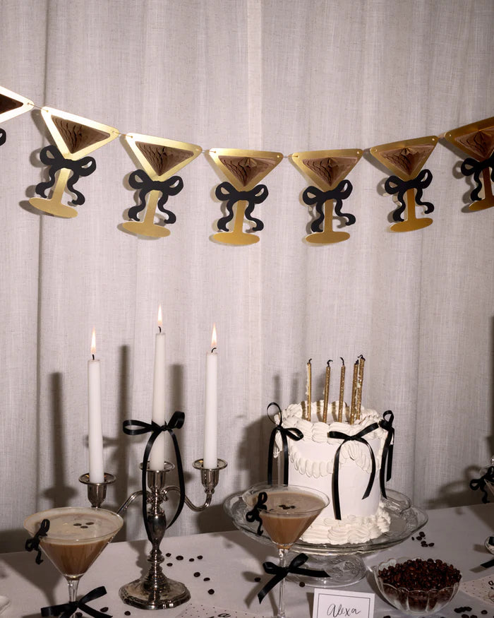 Martini glass bow banner hanging above espresso martini birthday dessert table with candlelit cake and cocktails