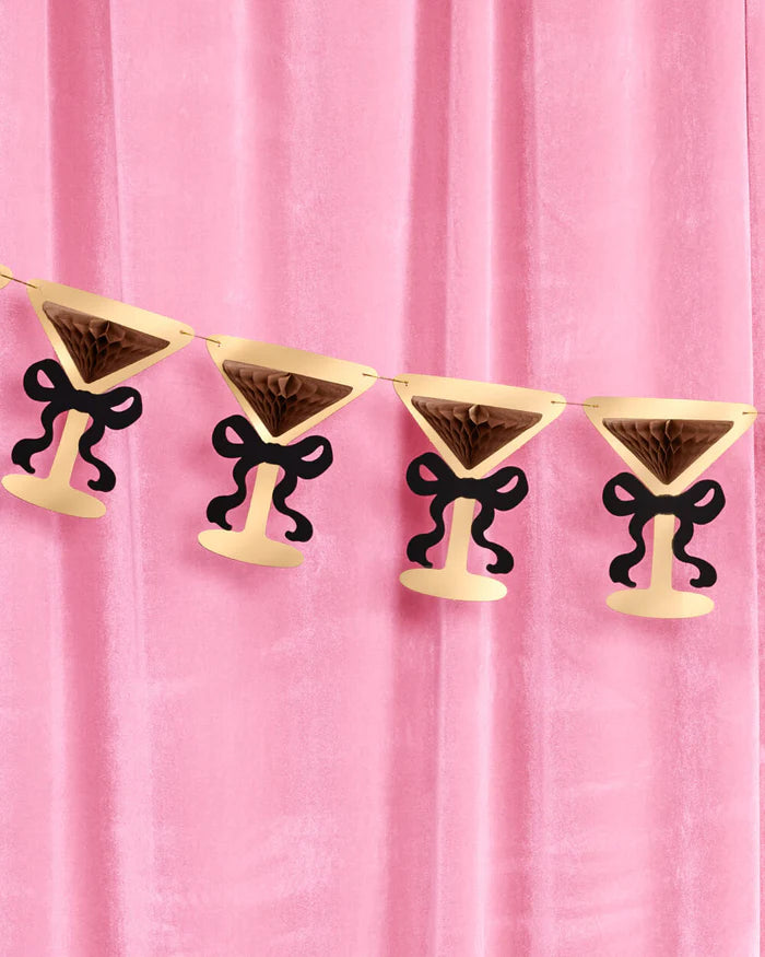 Gold foil martini glass bow banner hung against pink velvet backdrop with brown tissue accordions and black paper bows