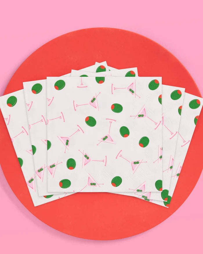 Fanned stack of martini and olive print cocktail napkins on a red plate with pink background