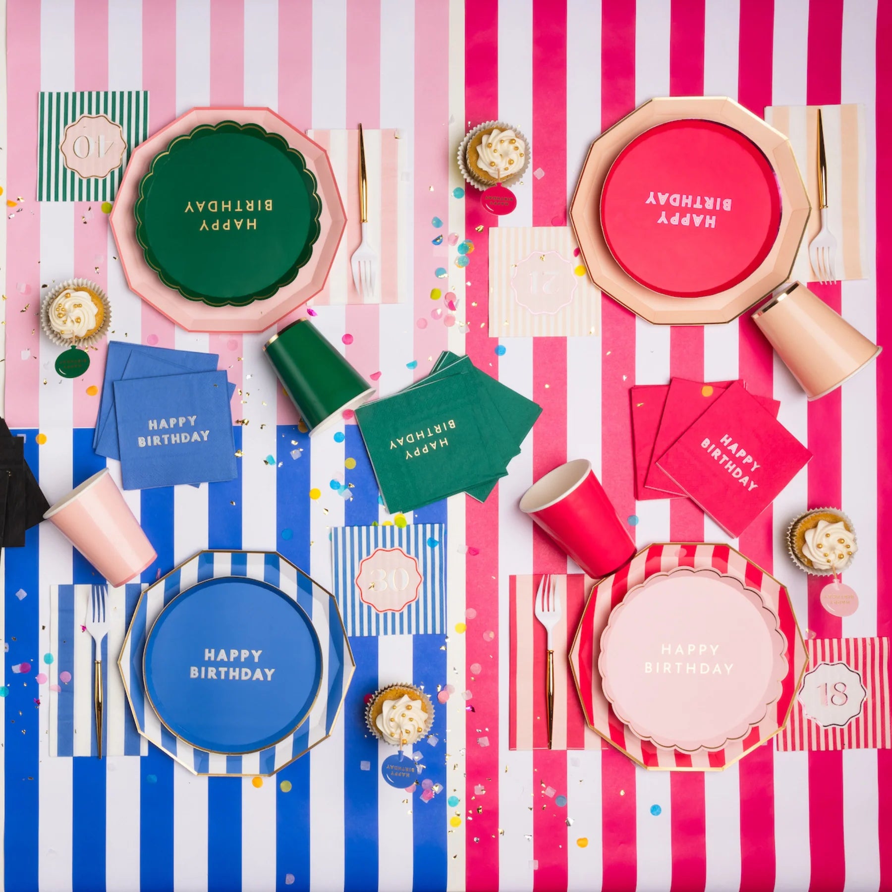 Overhead lifestyle shot of colorful milestone birthday party table with assorted Happy Birthday plates, napkins and cups in blue, green and pink on striped tablecloths with confetti