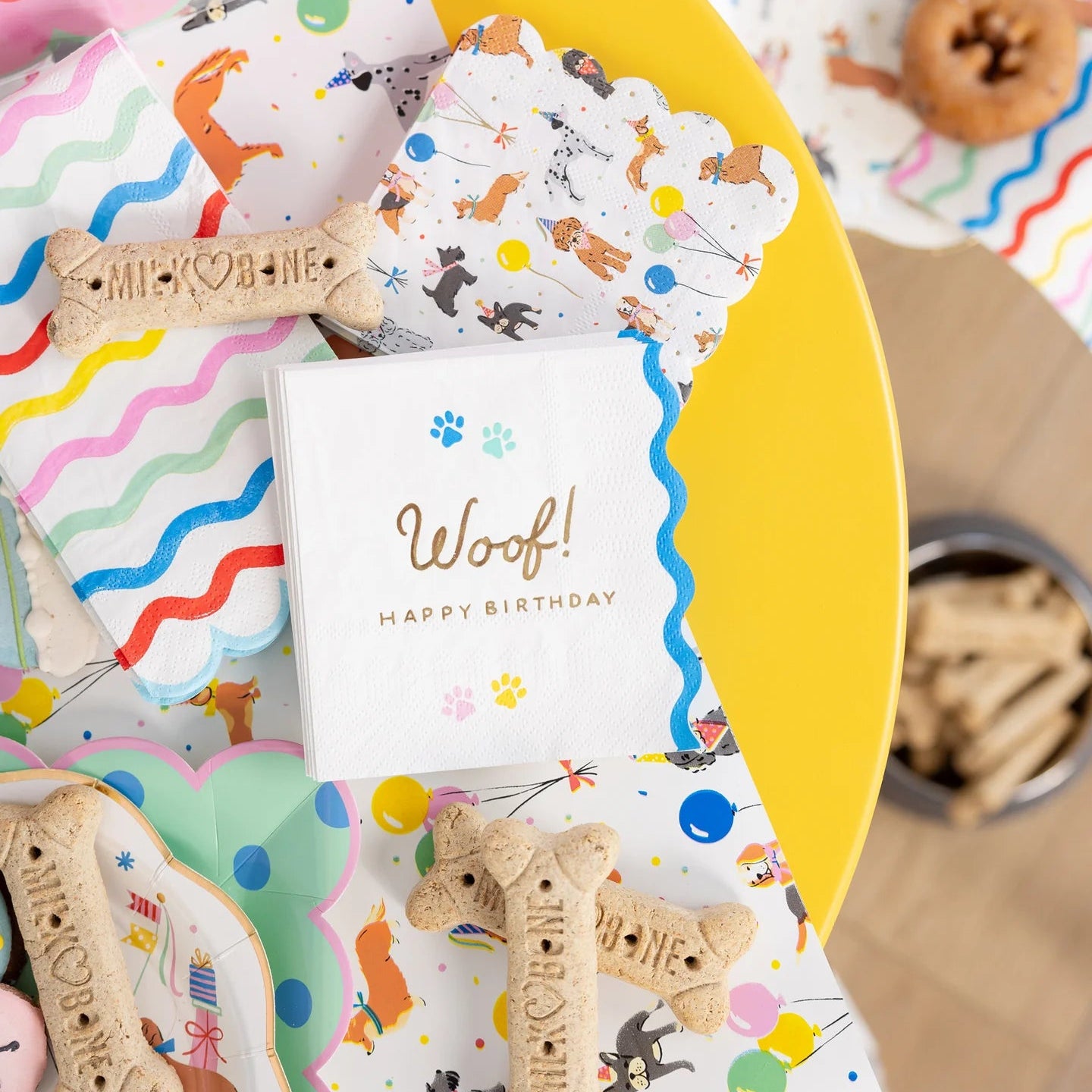 Party scene with dog napkins, bone treats, and rainbow stripe napkins on a yellow table.