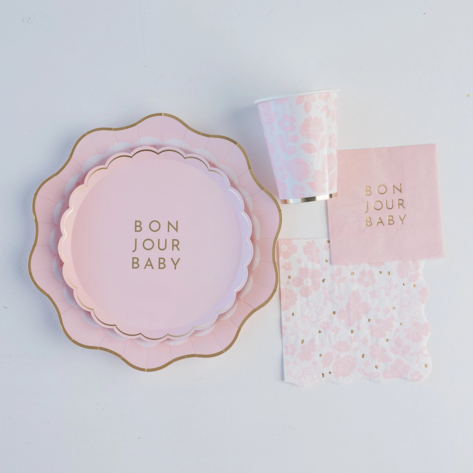 Flat‑lay of coordinating pink plates, floral cup and Bonjour Baby napkin for elegant baby shower place setting.