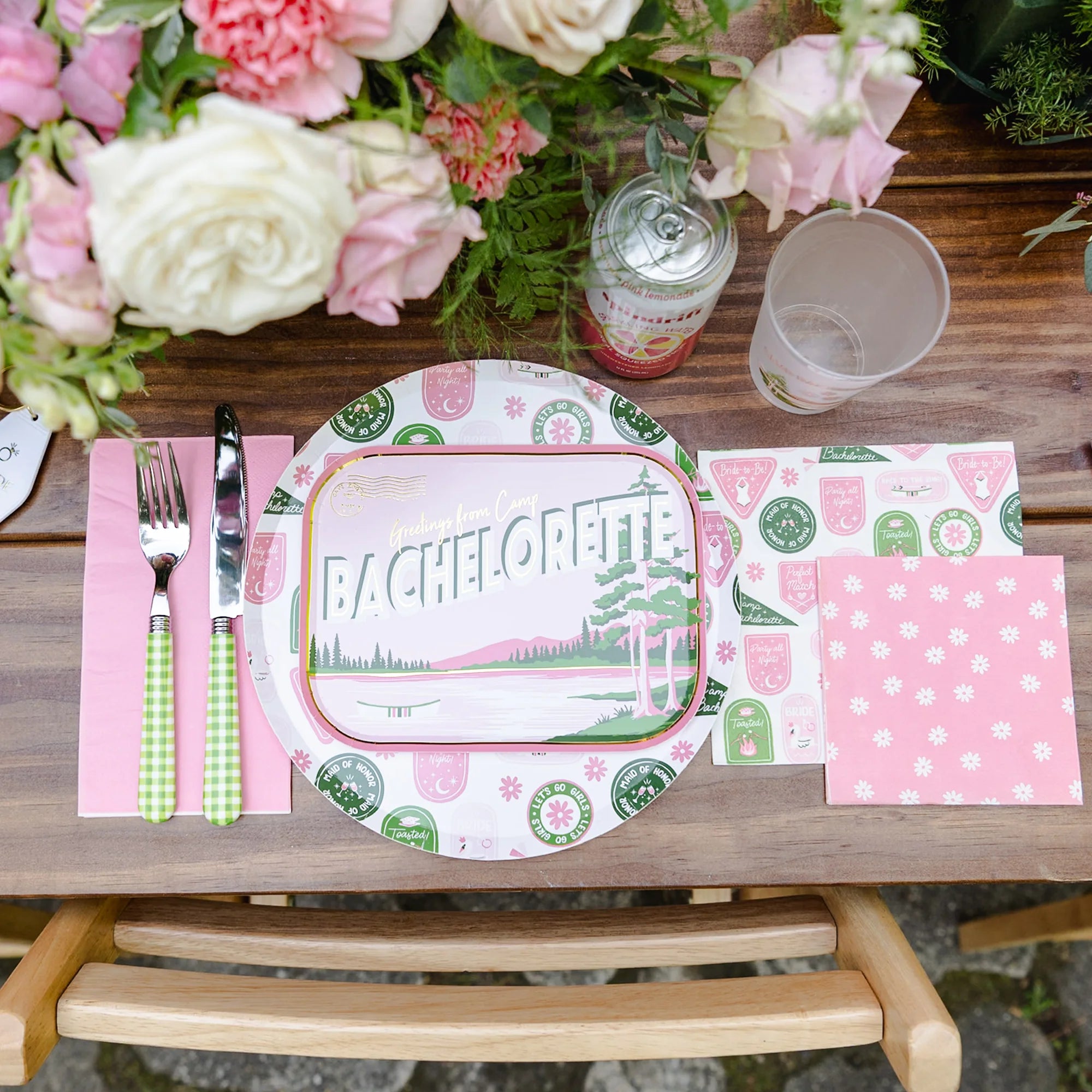Rustic outdoor tablescape with Camp Bachelorette plates and pink daisy cocktail napkins beside floral centerpieces
