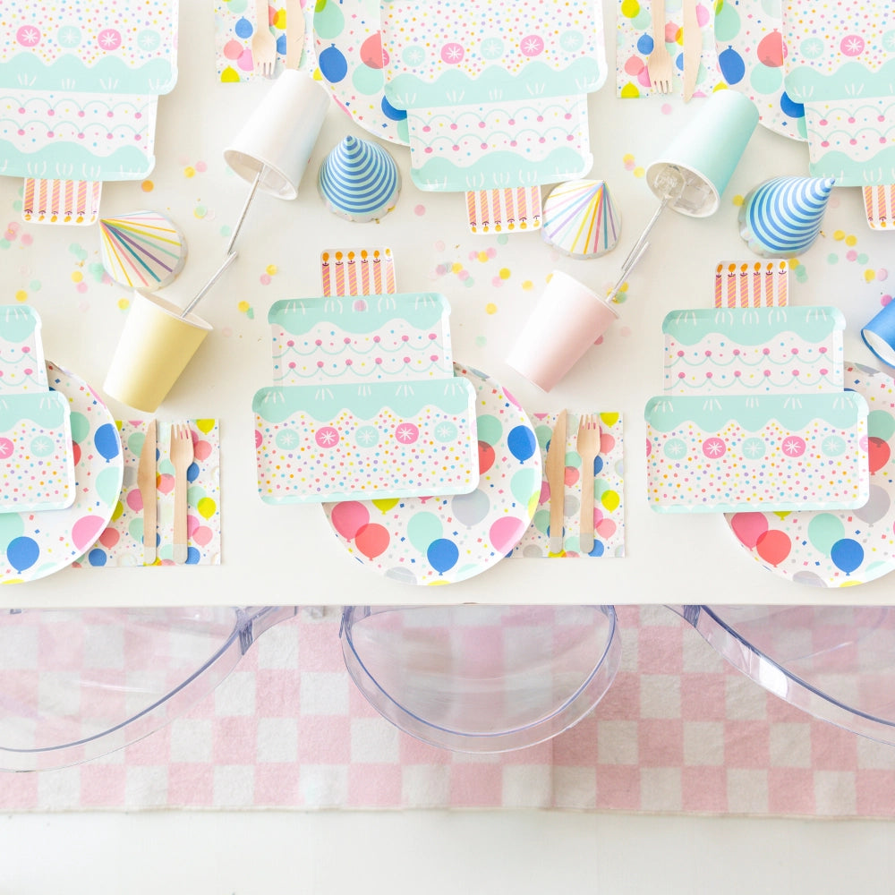 Overhead scene showing multiple cake plates, balloon plates, confetti napkins, and party hats on white table