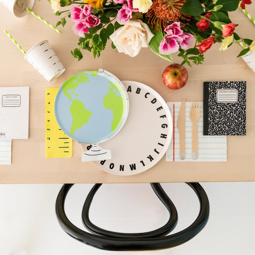 Overhead party setup: globe plate, alphabet charger, and ruler napkin under cup, surrounded by flowers and composition notebooks