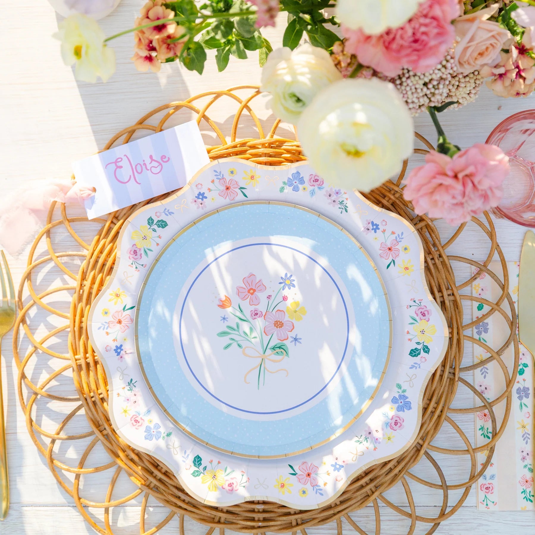 Overhead view of elegant floral scalloped dinner plate styled on a spring Easter table with pink roses, rattan charger, and place card