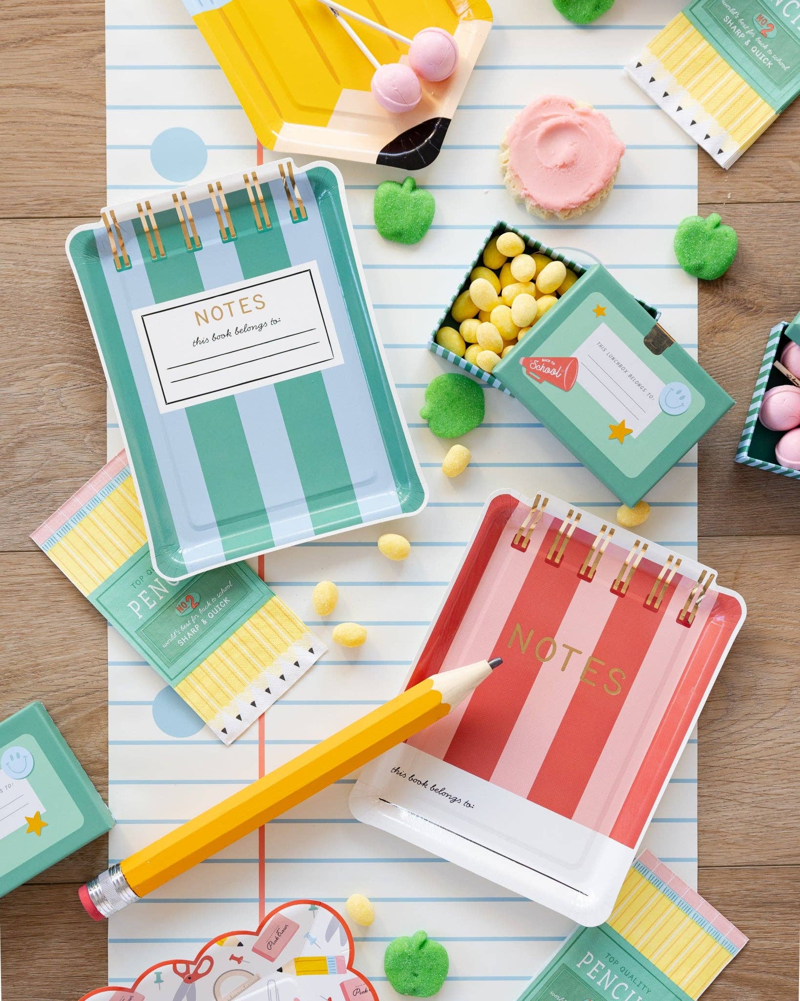 Flat lay of both notebook shaped paper plate designs — teal stripes and red stripes — styled with oversized pencil, pencil napkins, and school-themed treat boxes on notebook paper background