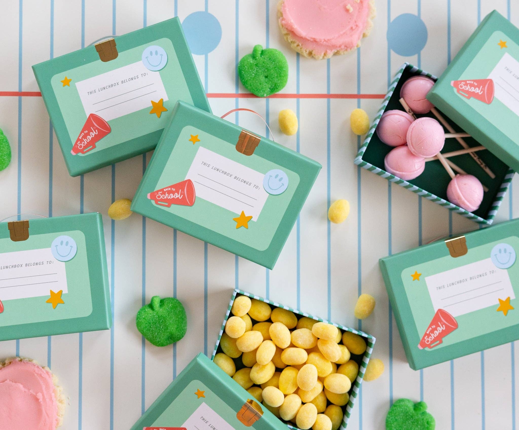 Flatlay of lunchbox shaped treat boxes styled on notebook paper table runner with green apple candies, pink frosted cookies, and yellow jelly candies