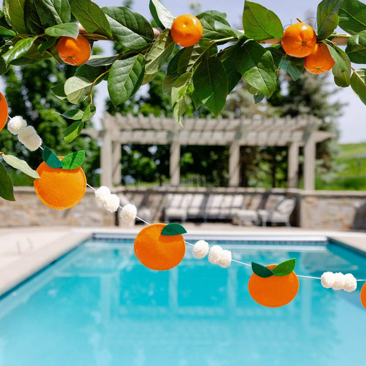 Orange felt garland hanging above a pool with fresh oranges and greenery.