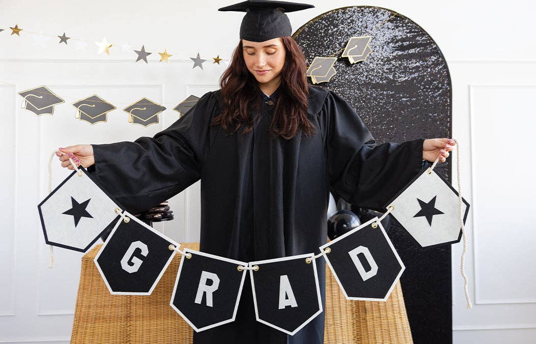 A graduate in a black gown holding a large graduation banner that spells GRAD with star accents.