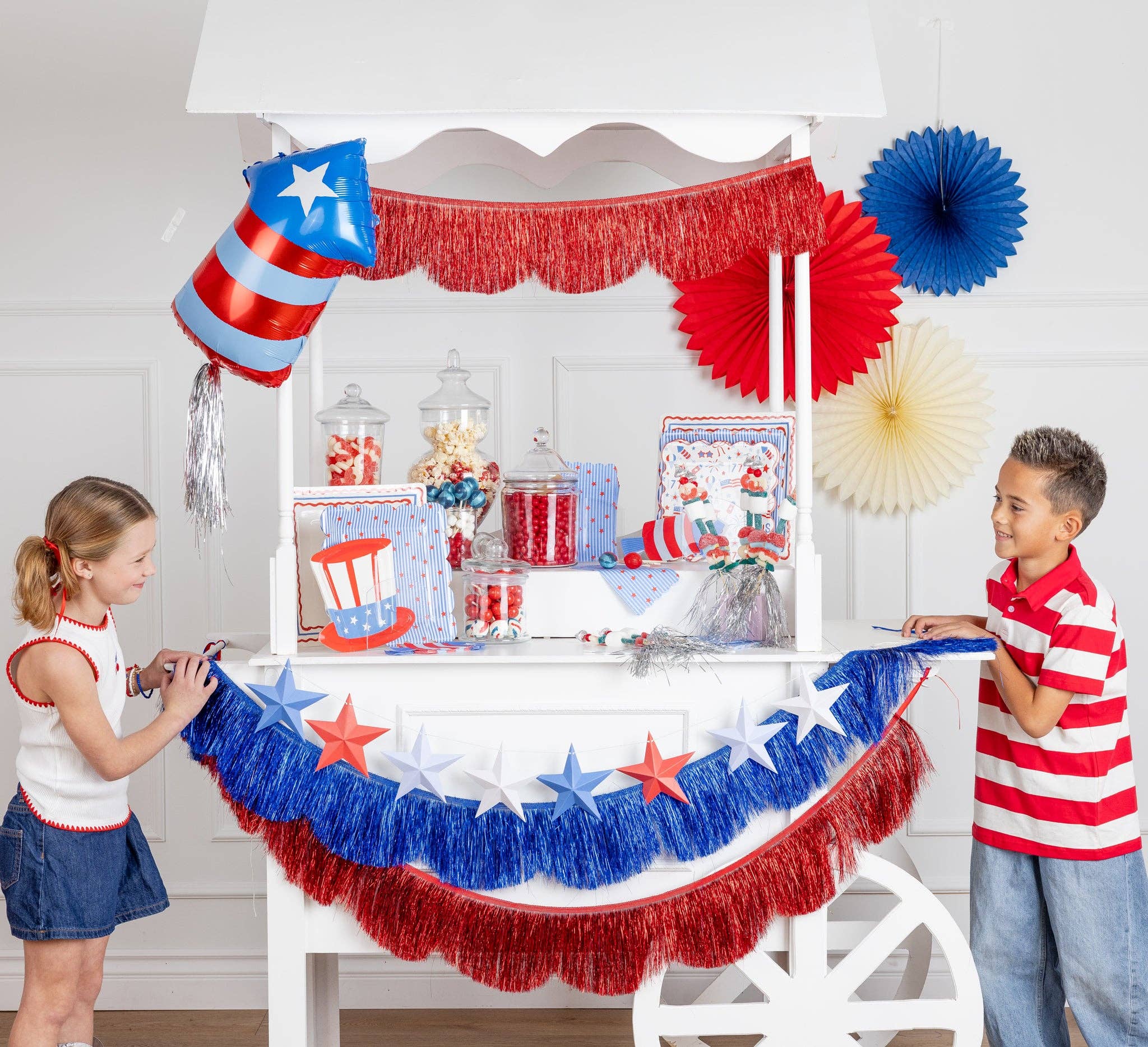 Two children holding a patriotic 3D paper star banner across a white party cart decorated with red and blue fringe garlands, a firecracker balloon, and paper fans