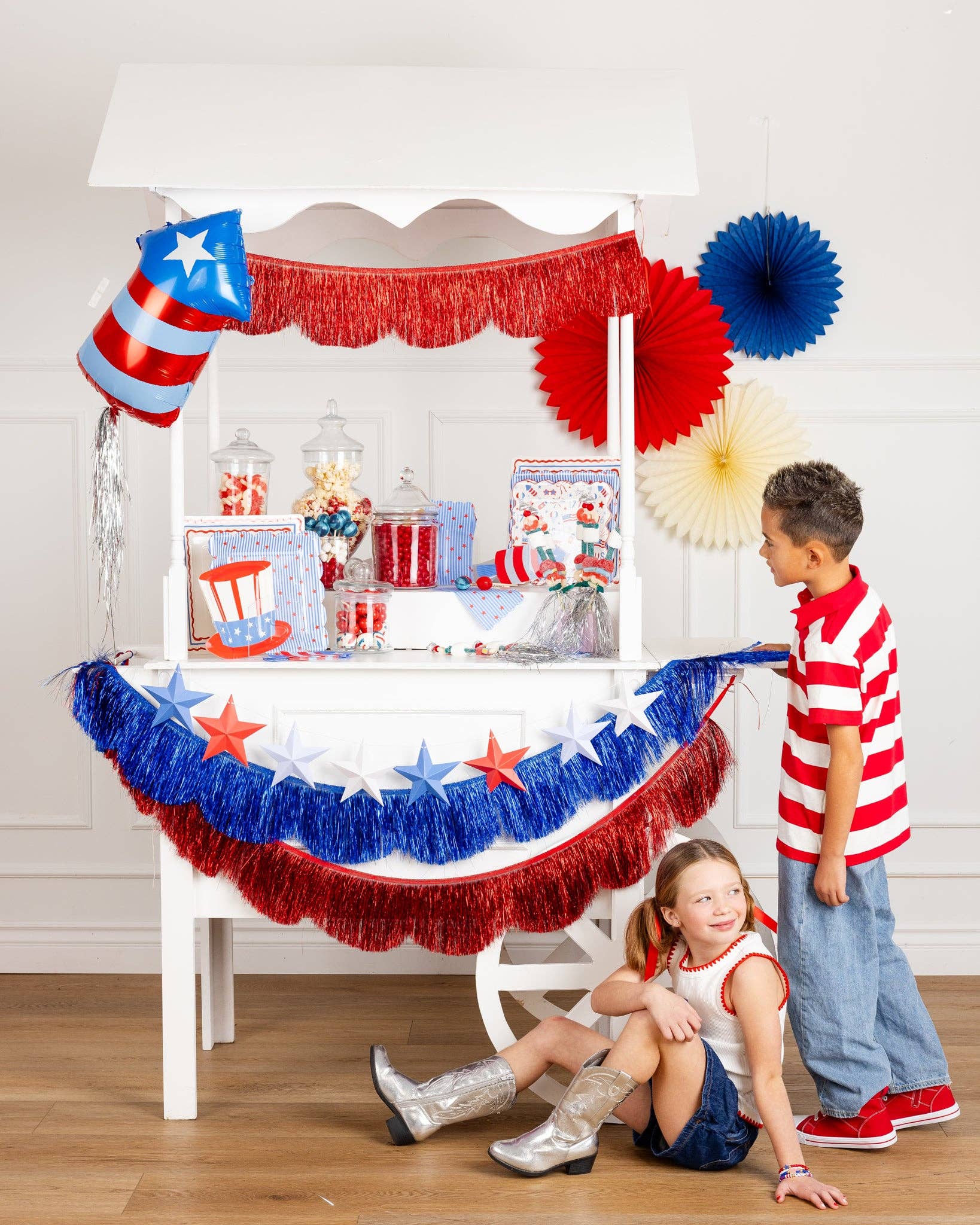 White party cart decorated for a 4th of July celebration with red and blue fringe banners, 3D star garland, a firecracker-shaped foil balloon, paper fans, and an assortment of patriotic paper plates, napkins, and candy jars, with two children standing nearby