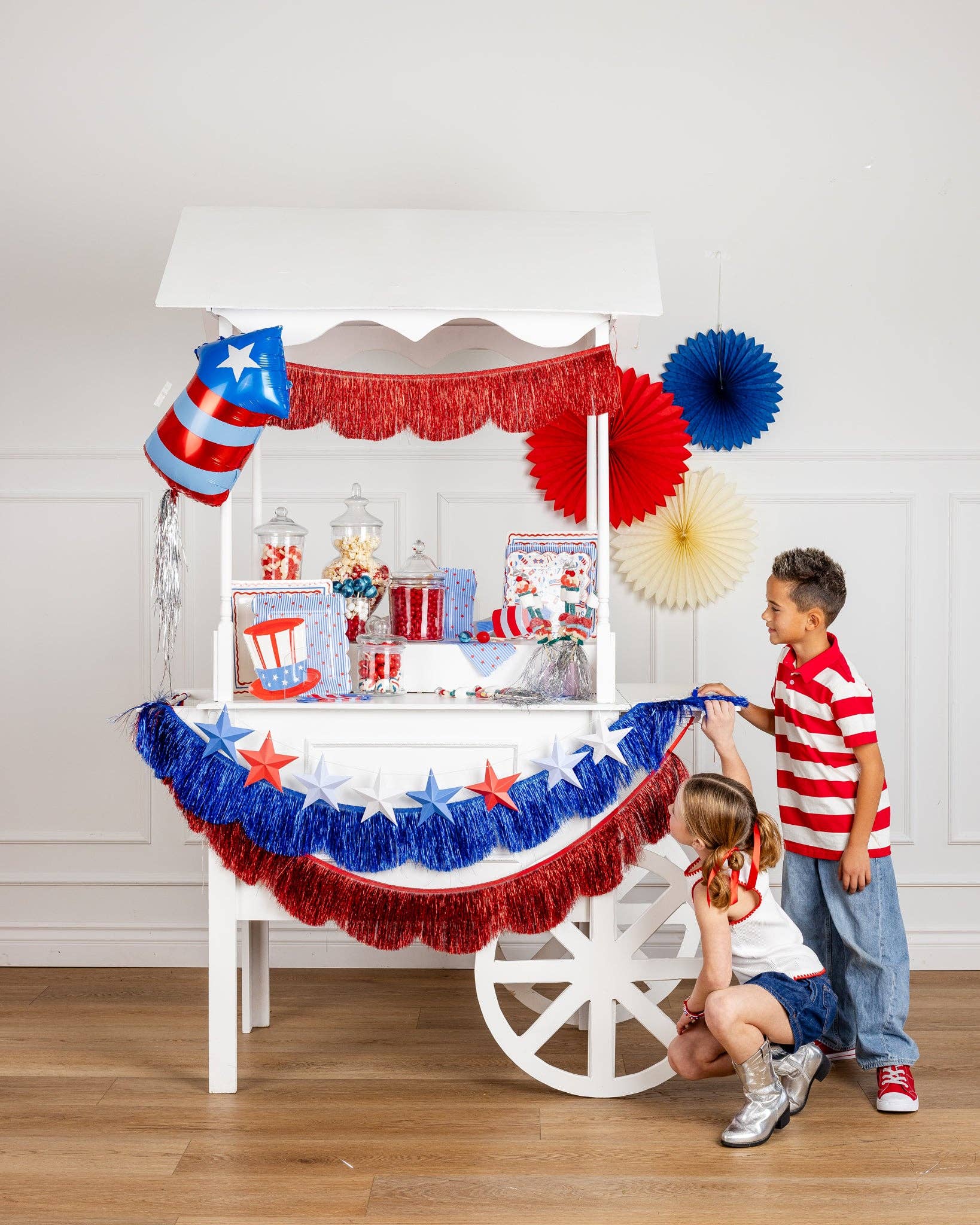 Patriotic candy cart with red white and blue tinsel fringe garlands star banner paper fans and glass jars of candy with two children in red white and blue outfits