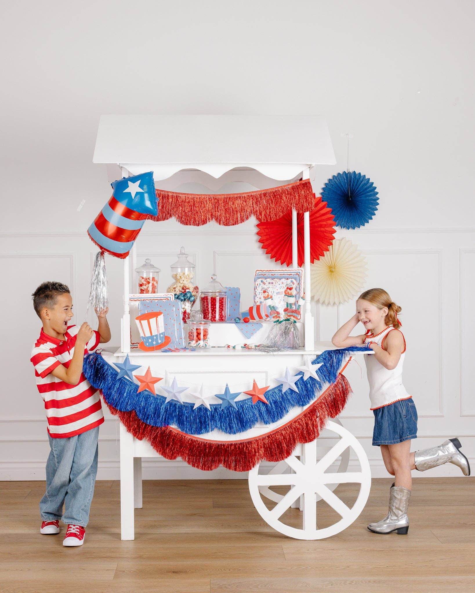 Two laughing children at a white party cart with a patriotic firecracker shaped mylar balloon, 3D star garland, and red and blue fringe banners