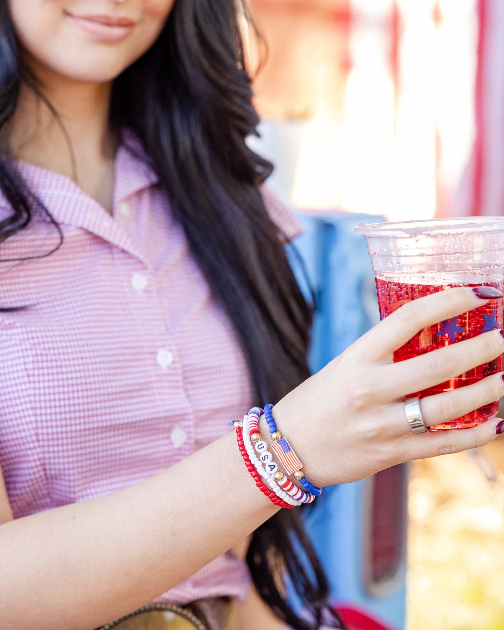 Woman holding a red drink and wearing a stack of four patriotic beaded bracelets with American flag charm and USA letter beads