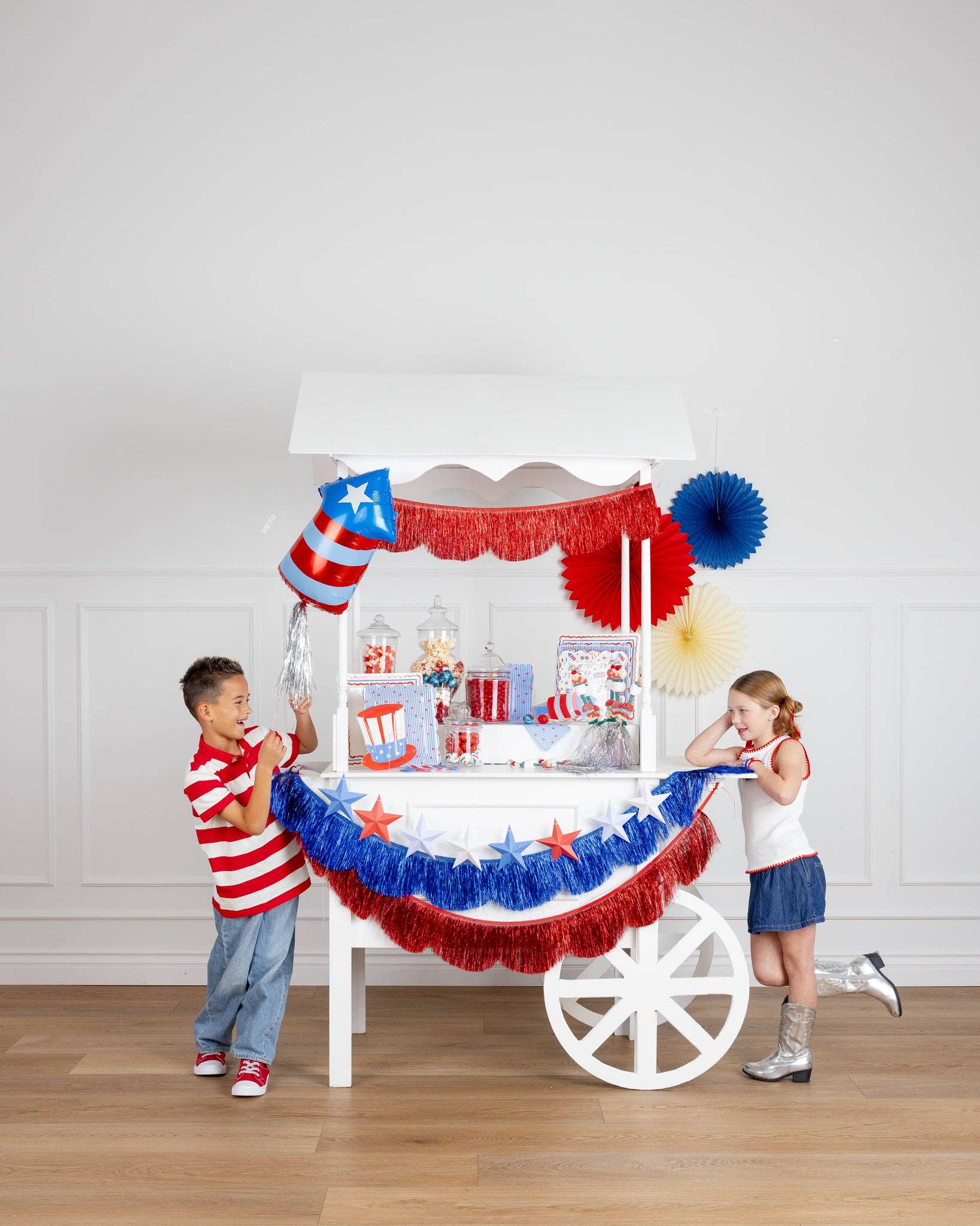 Two children at a white party cart decorated with red and blue metallic fringe scallop banners, a 3D paper star garland, firecracker balloon, paper fans, and patriotic tableware