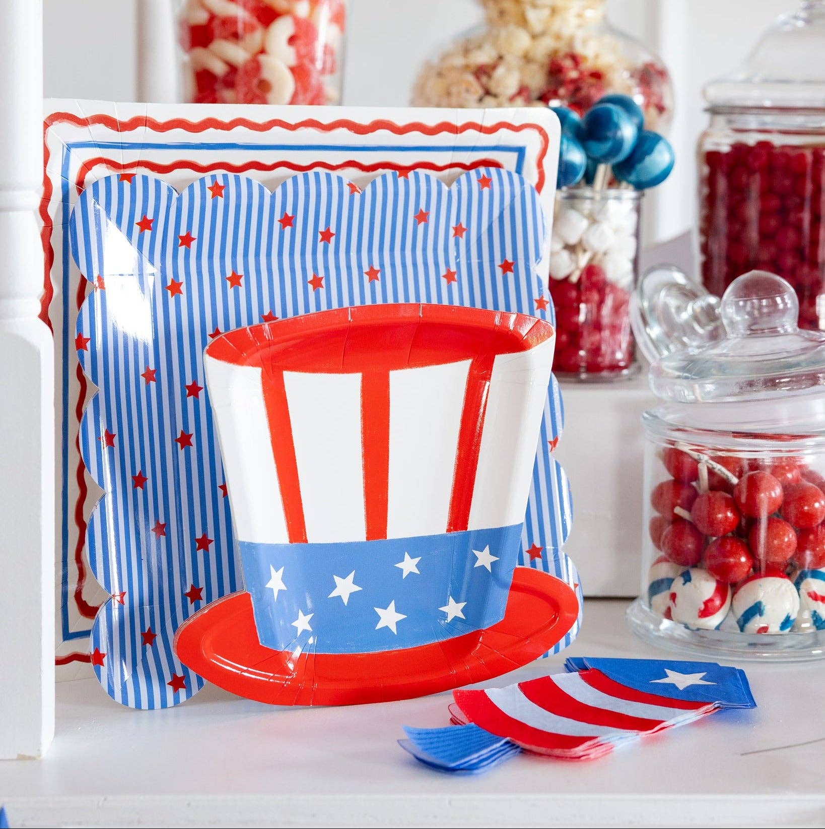 Patriotic paper plates stacked together on a candy table showing an Uncle Sam top hat shaped plate in front of a blue striped scalloped plate layered over a cream square border dinner plate, with firecracker shaped napkins and glass candy jars in the background