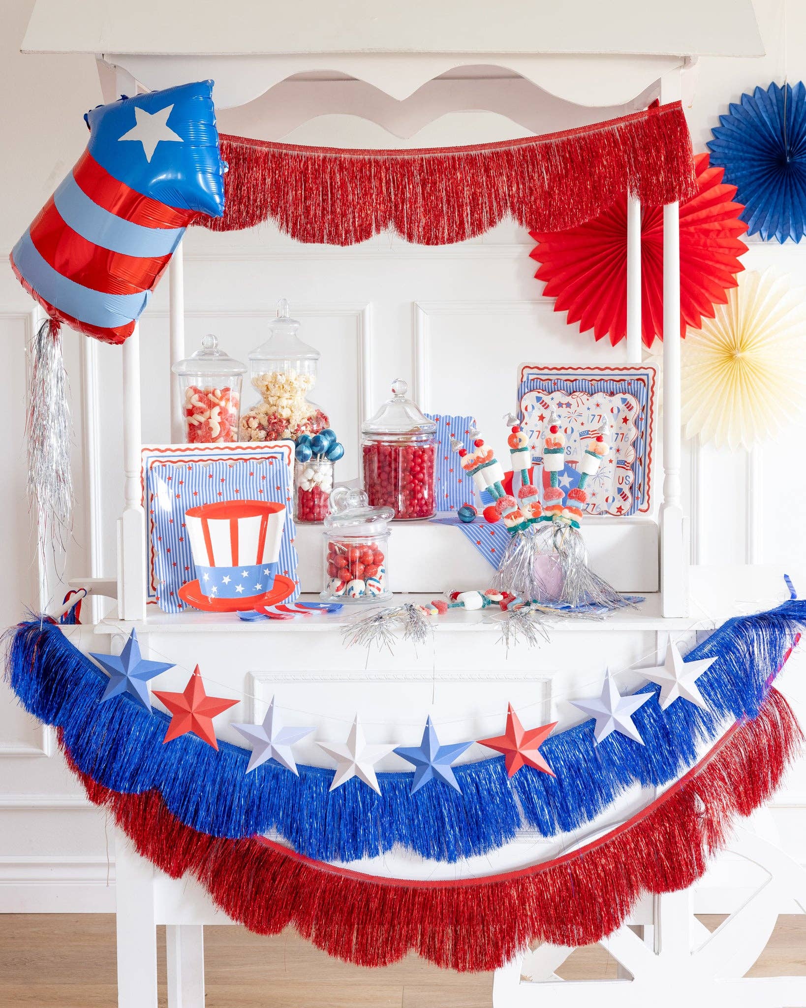 White party cart decorated for 4th of July with red and blue fringe scallop banners, 3D paper star garland, firecracker balloon, paper fans, and patriotic tableware with candy jars