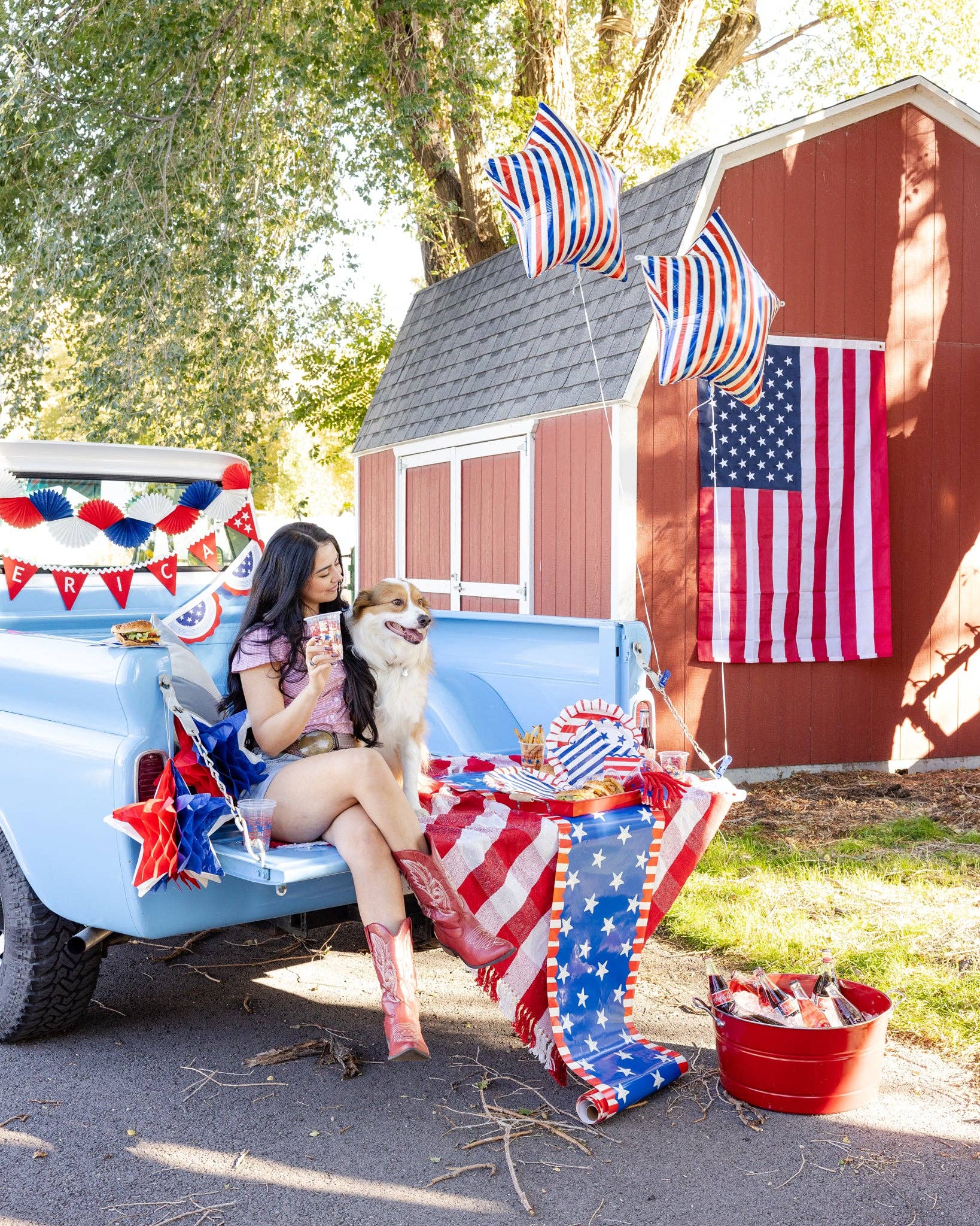 Wide lifestyle shot of a patriotic truck bed party setup with woman holding a stars and stripes clear plastic cup alongside coordinating plates and banners