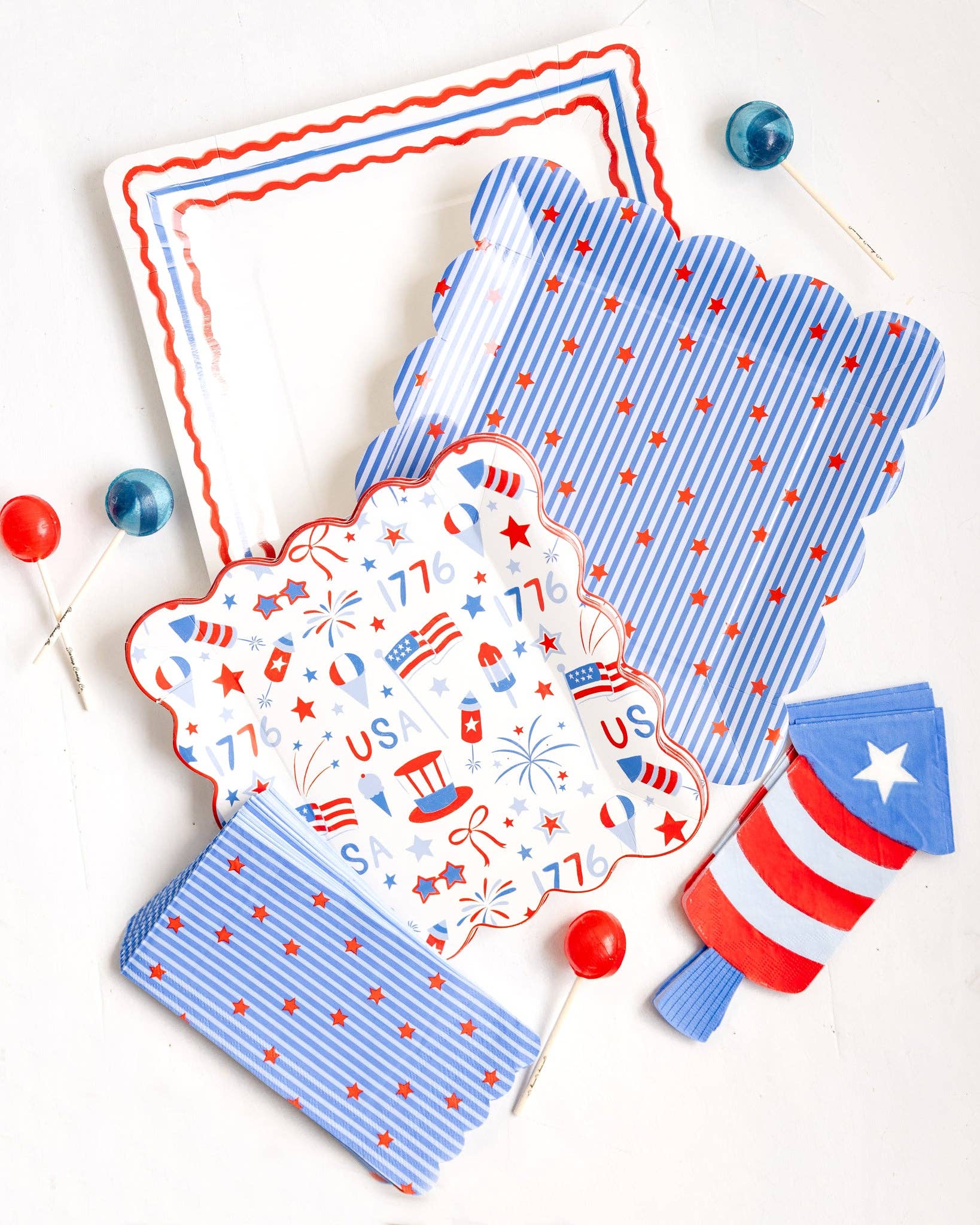 Flat lay of full patriotic plate and napkin collection showing stars and stripes square plates 1776 USA scalloped plates and firecracker shaped napkins with cake pops