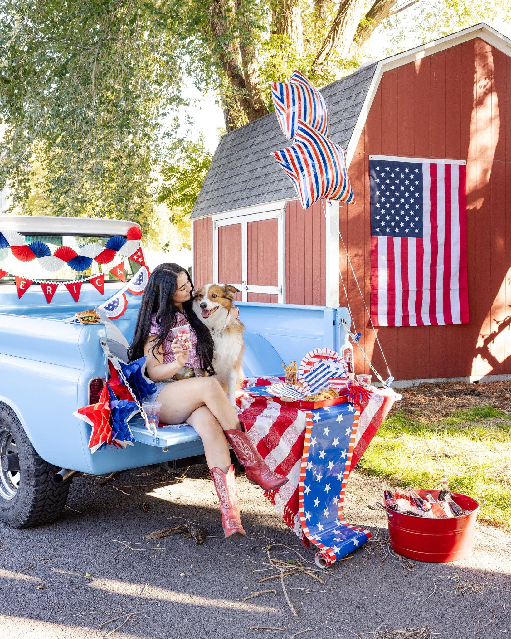 Patriotic tailgate party in a vintage blue truck bed with American flag, red white and blue paper fans, star balloon, striped plates, and picnic spread with burgers and drinks