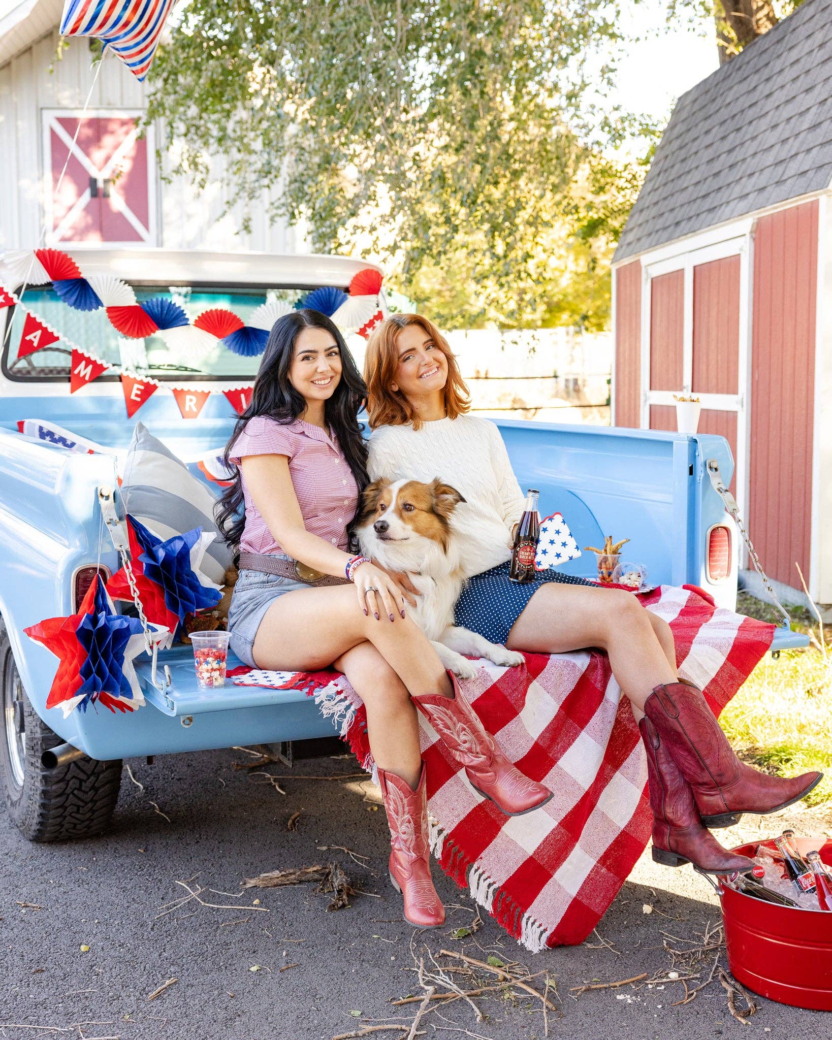 Two women sitting in decorated blue vintage truck bed with red white and blue tissue fan bunting banner and patriotic party decorations in background