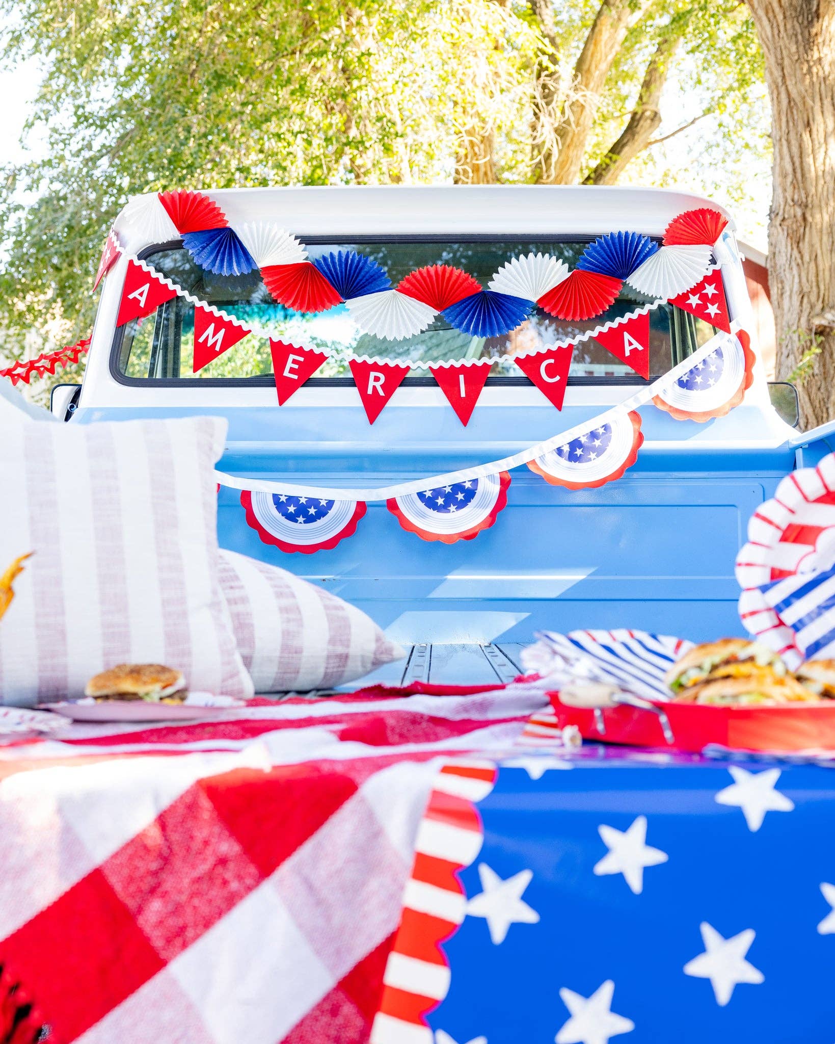 Red white and blue tissue fan bunting banner hung on vintage truck at a styled 4th of July picnic with American flag table runner and party supplies