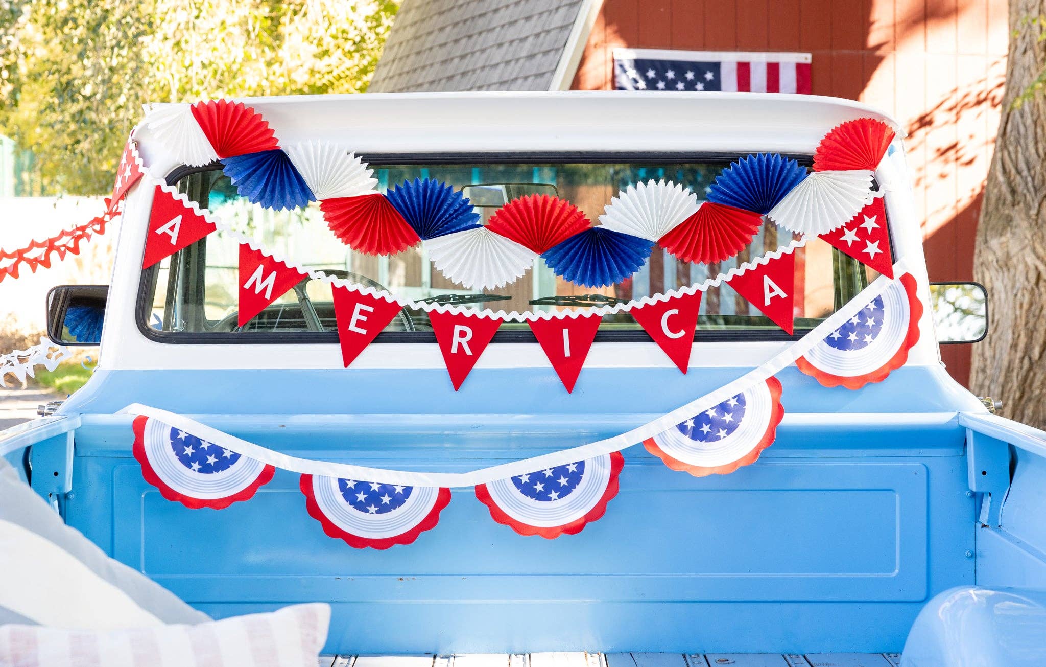 Red white and blue tissue fan bunting banner hung across vintage blue truck back window with America felt pennant banner and patriotic bunting garland at a 4th of July celebration