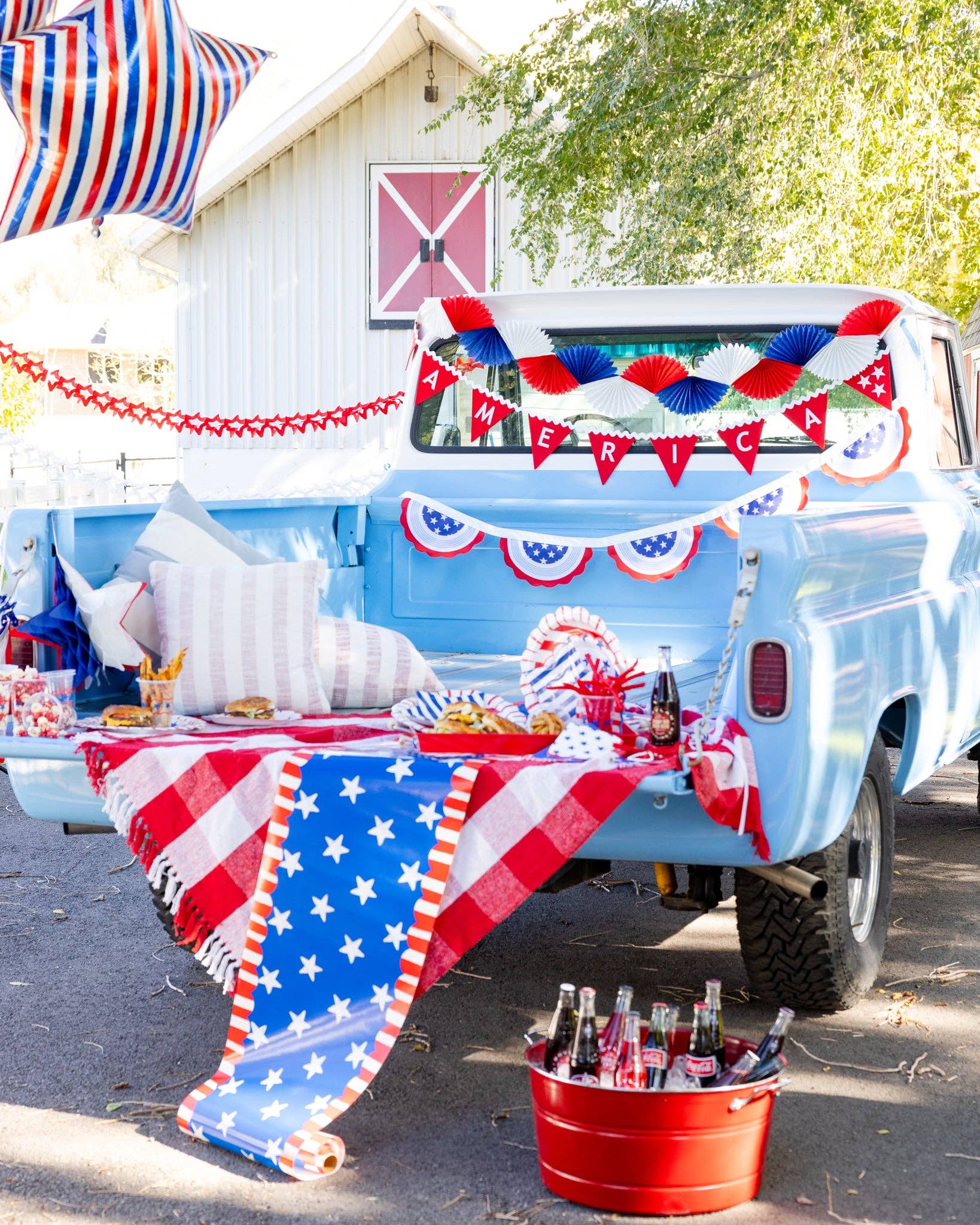 Red white and blue tissue fan bunting banner hung on blue vintage truck at 4th of July party with America felt pennant banner striped star balloon and patriotic decor