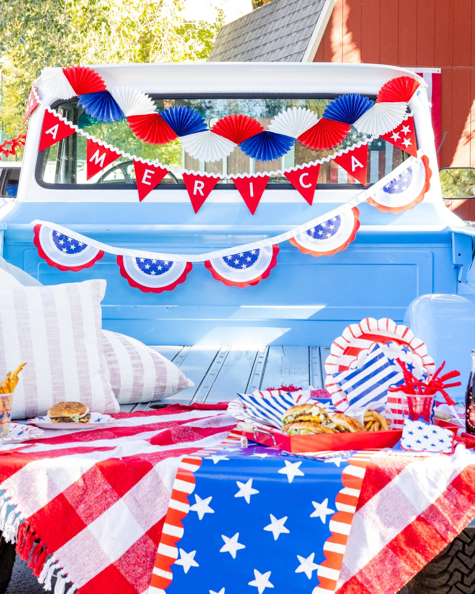 Patriotic truck bed party setup with red white and blue paper fans, AMERICA pennant banner, bunting garland, star-patterned tablecloth, striped plates, burgers, and fries styled for a Fourth of July celebration