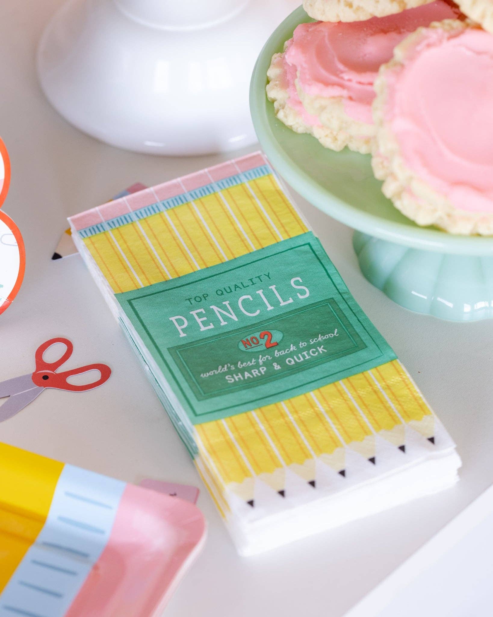 Pencil box paper dinner napkin styled on white table next to green cake stand with pink frosted cookies, scissors paper confetti, and pencil confetti