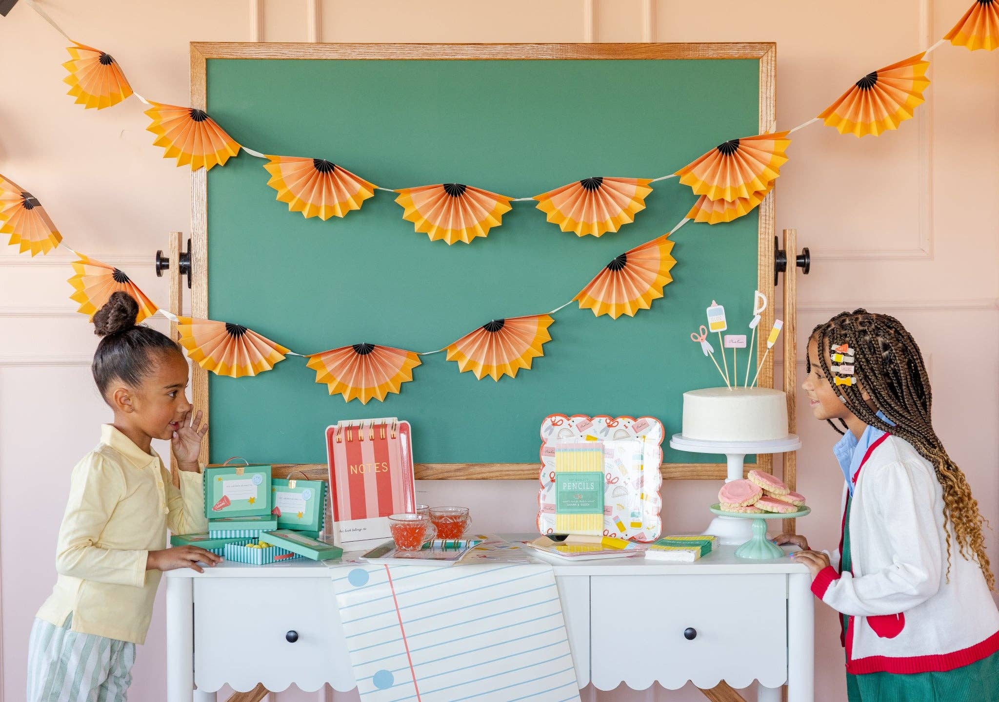 Two children at styled back to school party table with pencil shaving banner hanging above chalkboard, cake, and classroom-themed decor